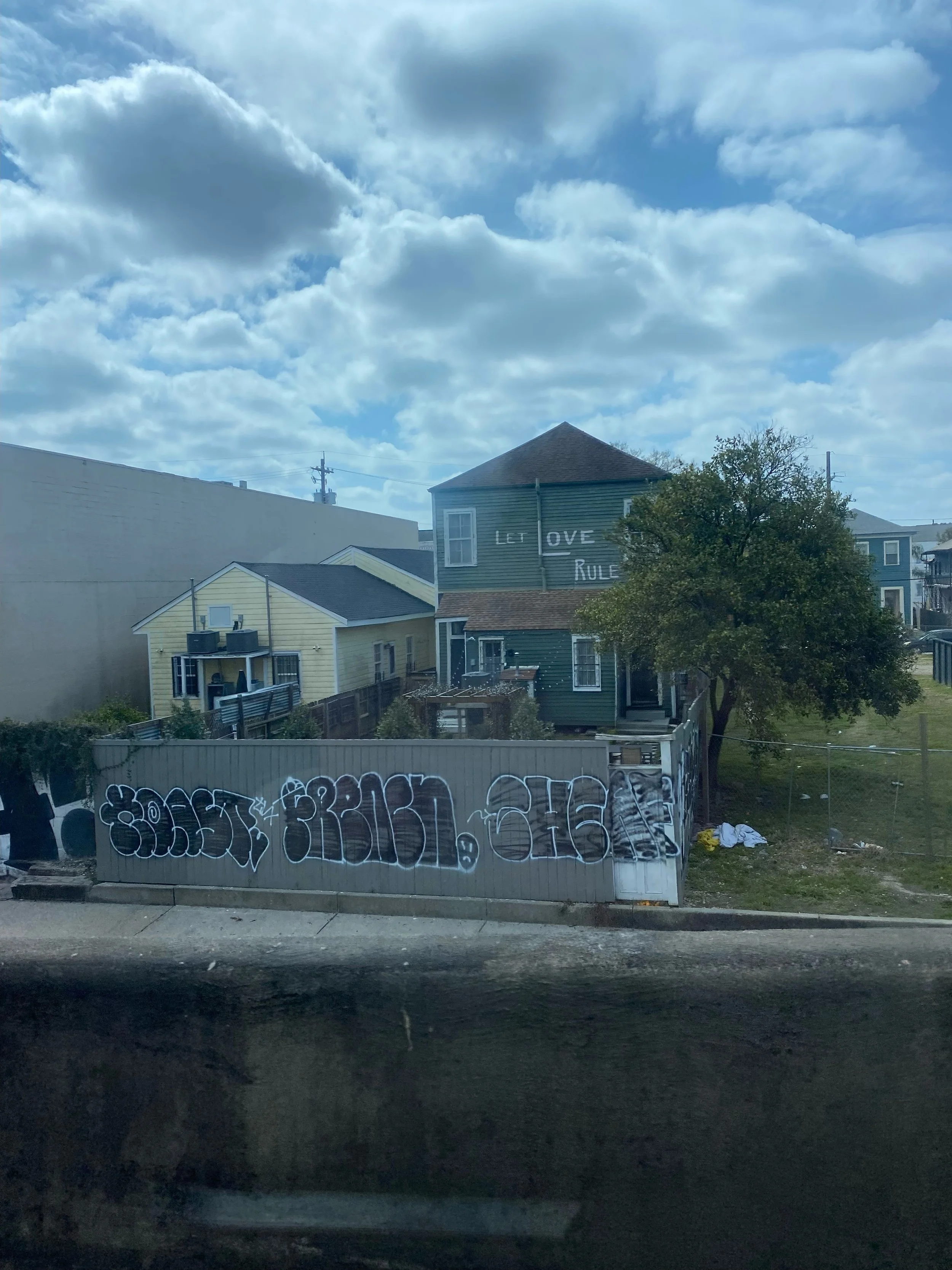 A residential neighborhood with houses, a tree, graffiti on a fence, and a cloudy sky. The house on the right has a sign that reads 'LET LOVE RULE' on its upper story.