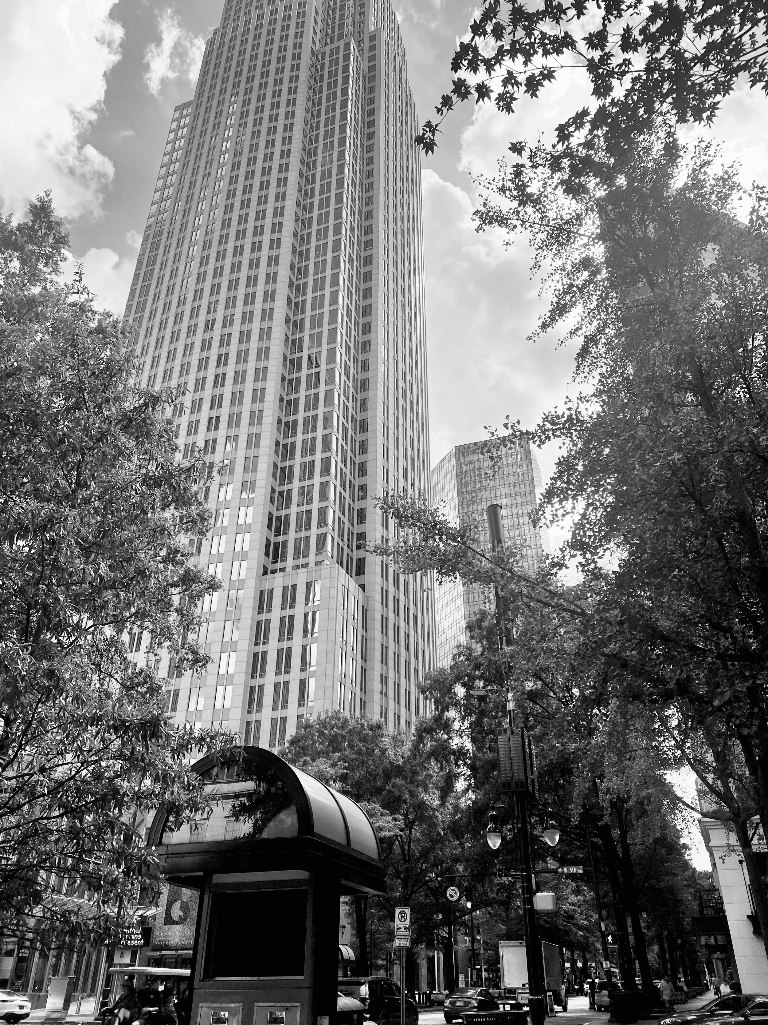 Black and white photo of a tall skyscraper with many windows, surrounded by trees and smaller buildings.