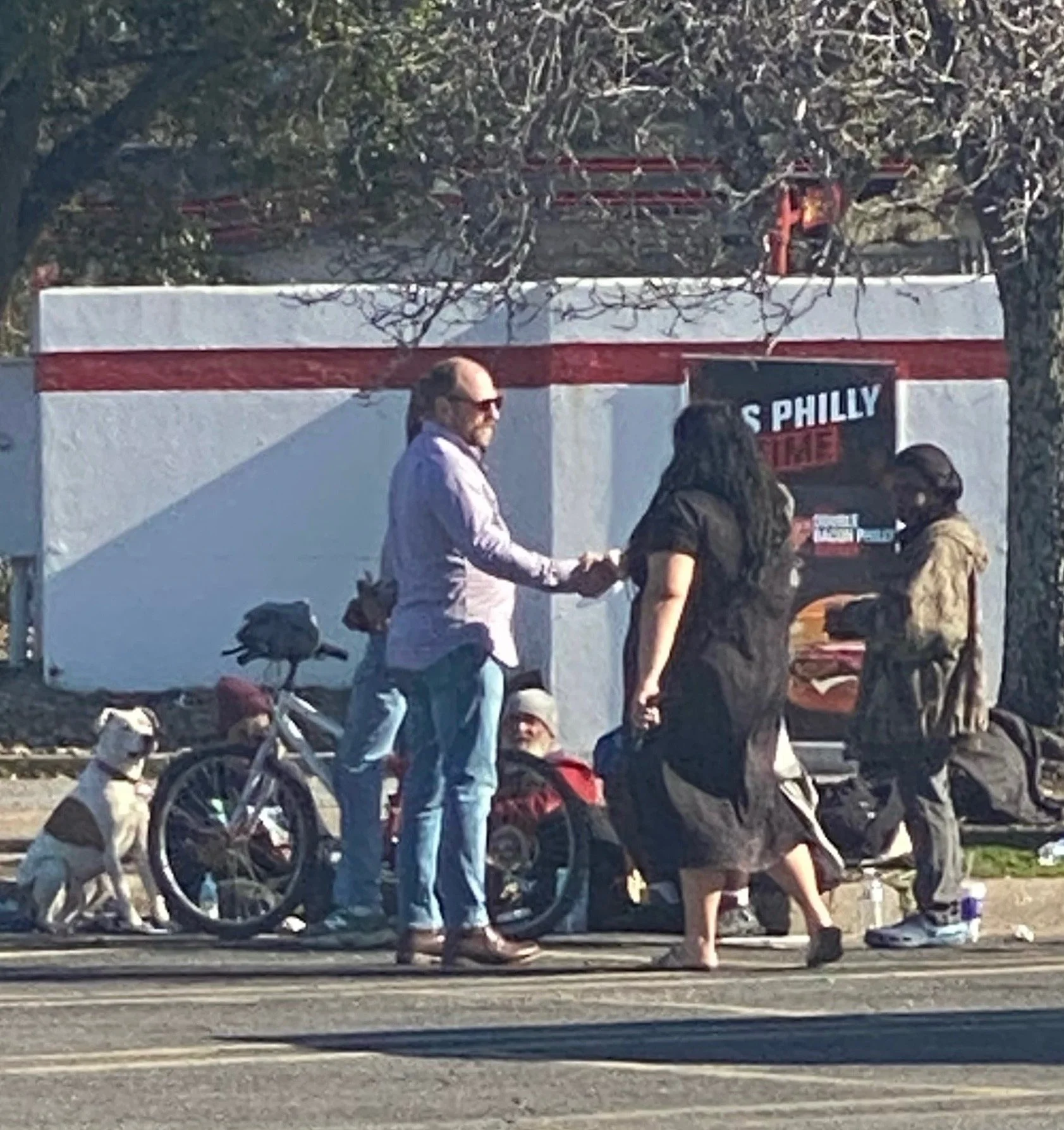 People standing on the sidewalk, talking, with a dog and bicycles nearby. A man in sunglasses and a woman are shaking hands. There are trees and a sign in the background.