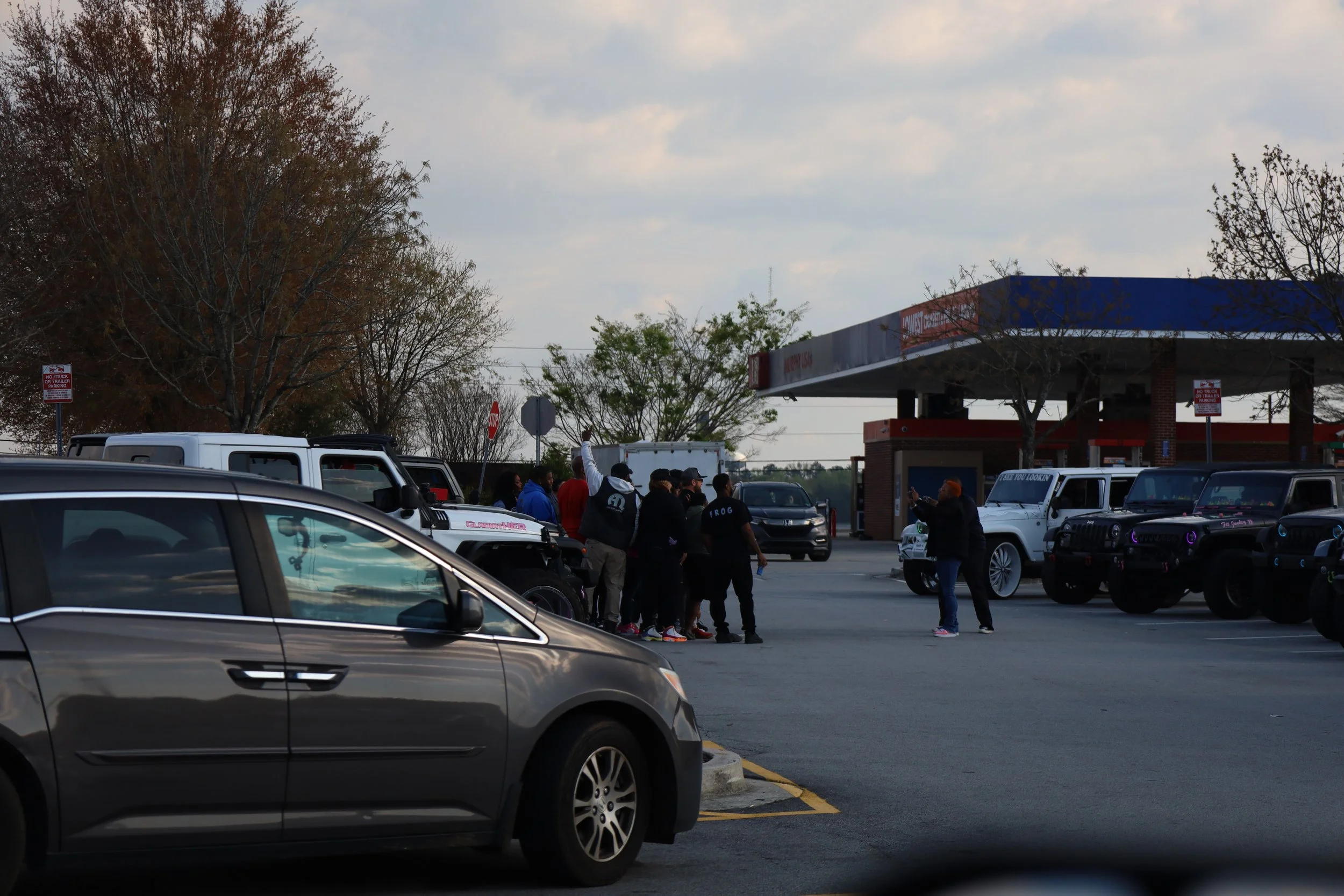 A group of people gathered outside a gas station, with some vehicles parked nearby during daytime.