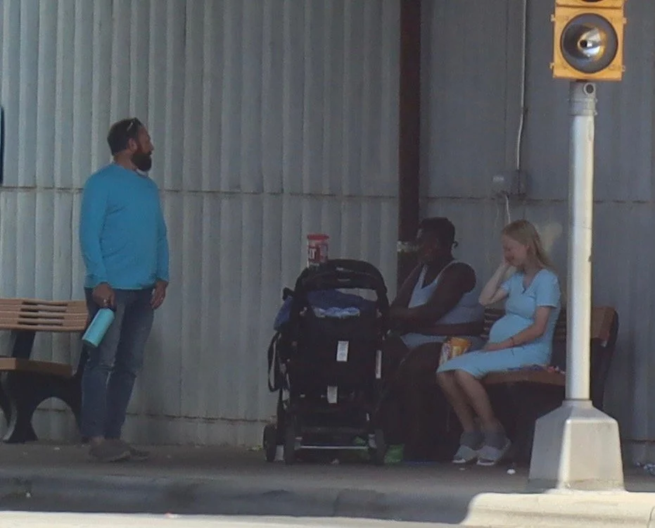 Three women sitting on a bench at a bus stop, one bound in a light blue dress, one with dark skin wearing a black top, and a pregnant woman with a light blue dress, with a man standing nearby holding a water bottle.