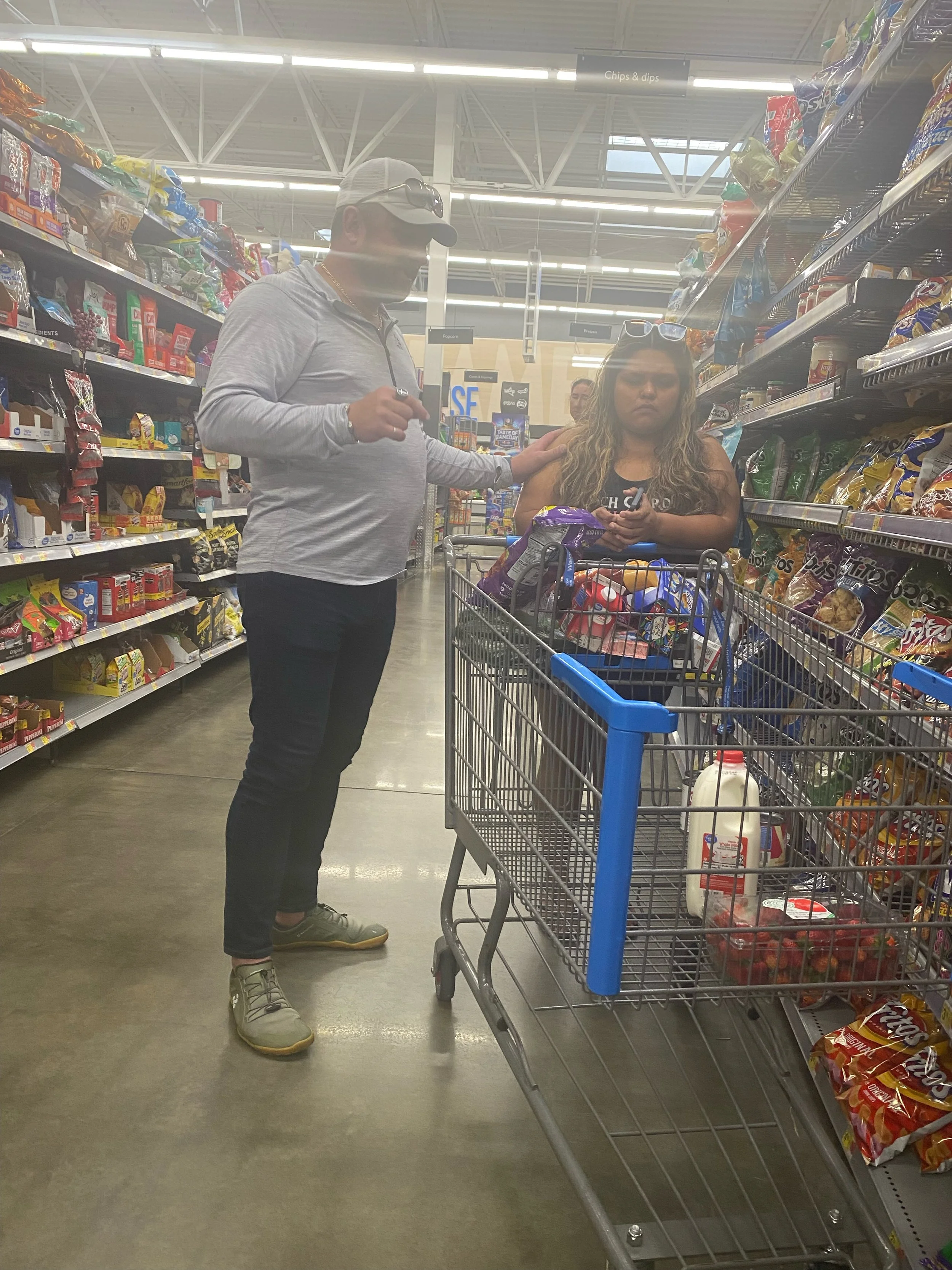 A man prays for a woman shopping in a grocery store aisle with snacks and chips. The man is wearing a gray long-sleeve shirt, dark pants, and a cap, and is touching the woman's shoulder. 
