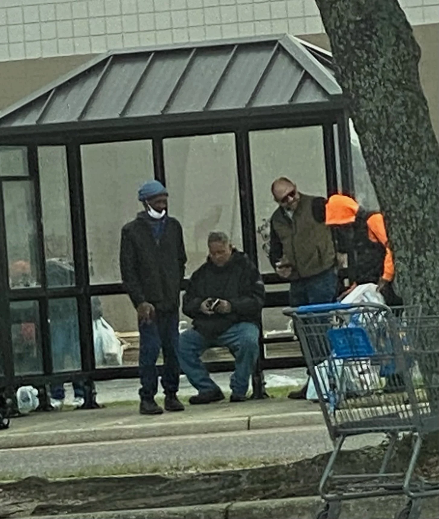 Four men standing and sitting outside a glass bus stop shelter, with a shopping cart nearby. One man is seated, others are standing, all appear engaged in conversation and prayer.