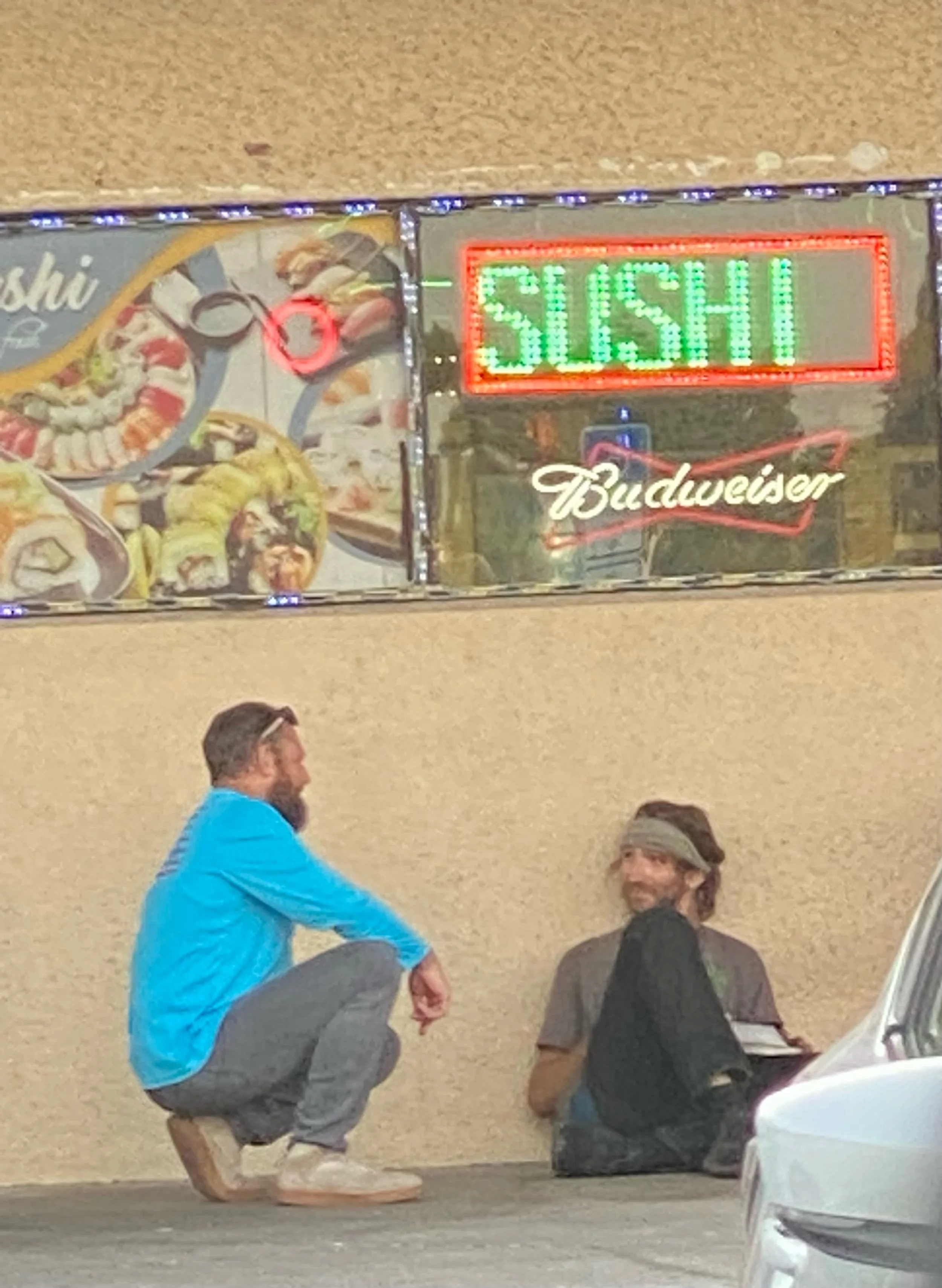 Two men sitting on the sidewalk in front of a sushi restaurant, one crouched down and one sitting on the ground, with a neon sign reading 'SUSHI' and a picture of sushi dishes above them.