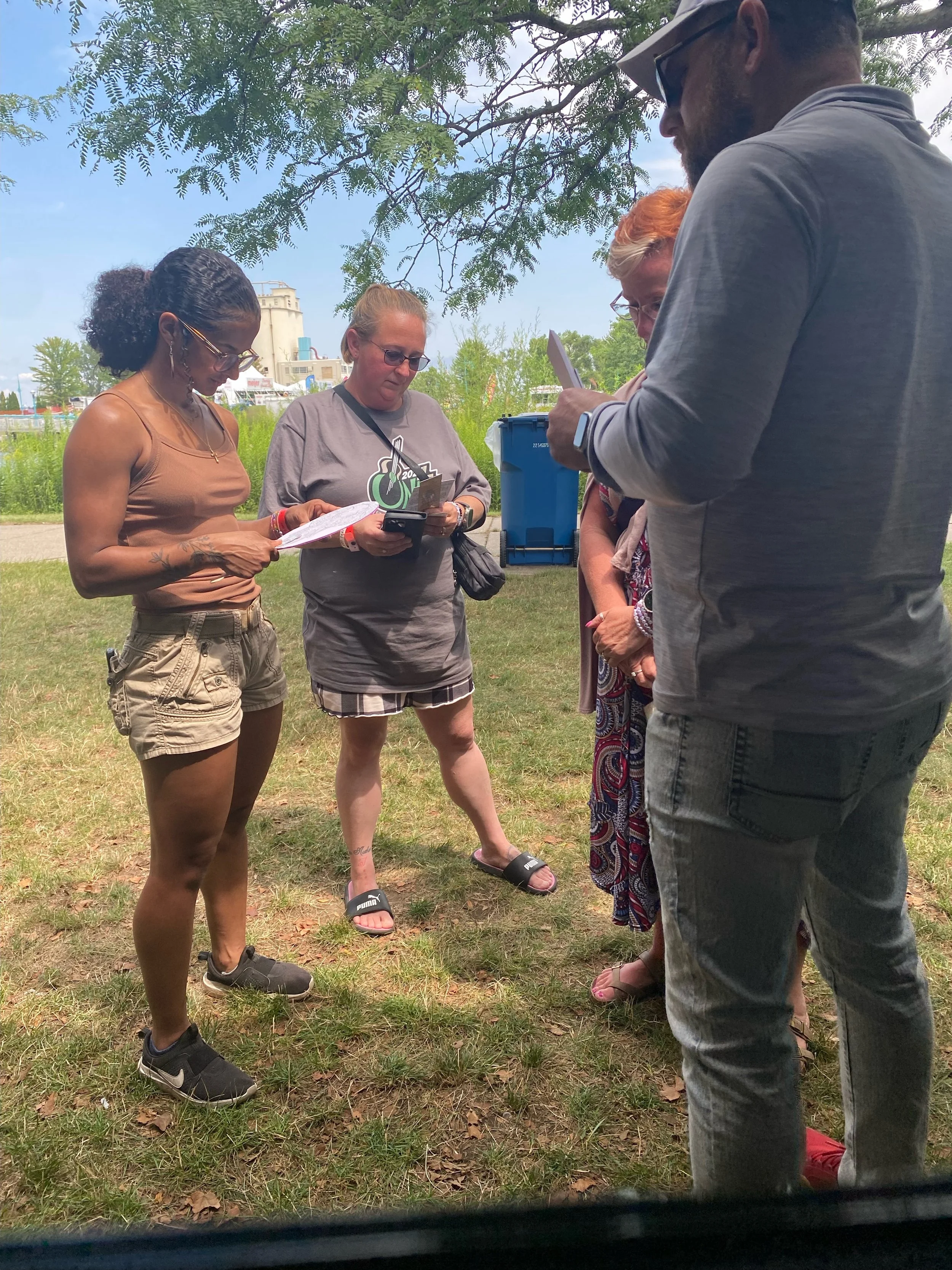 Group of six people standing outdoors under a tree, looking at papers and a phone, with a background of green grass, trees, a blue recycling bin, and a building in the distance.