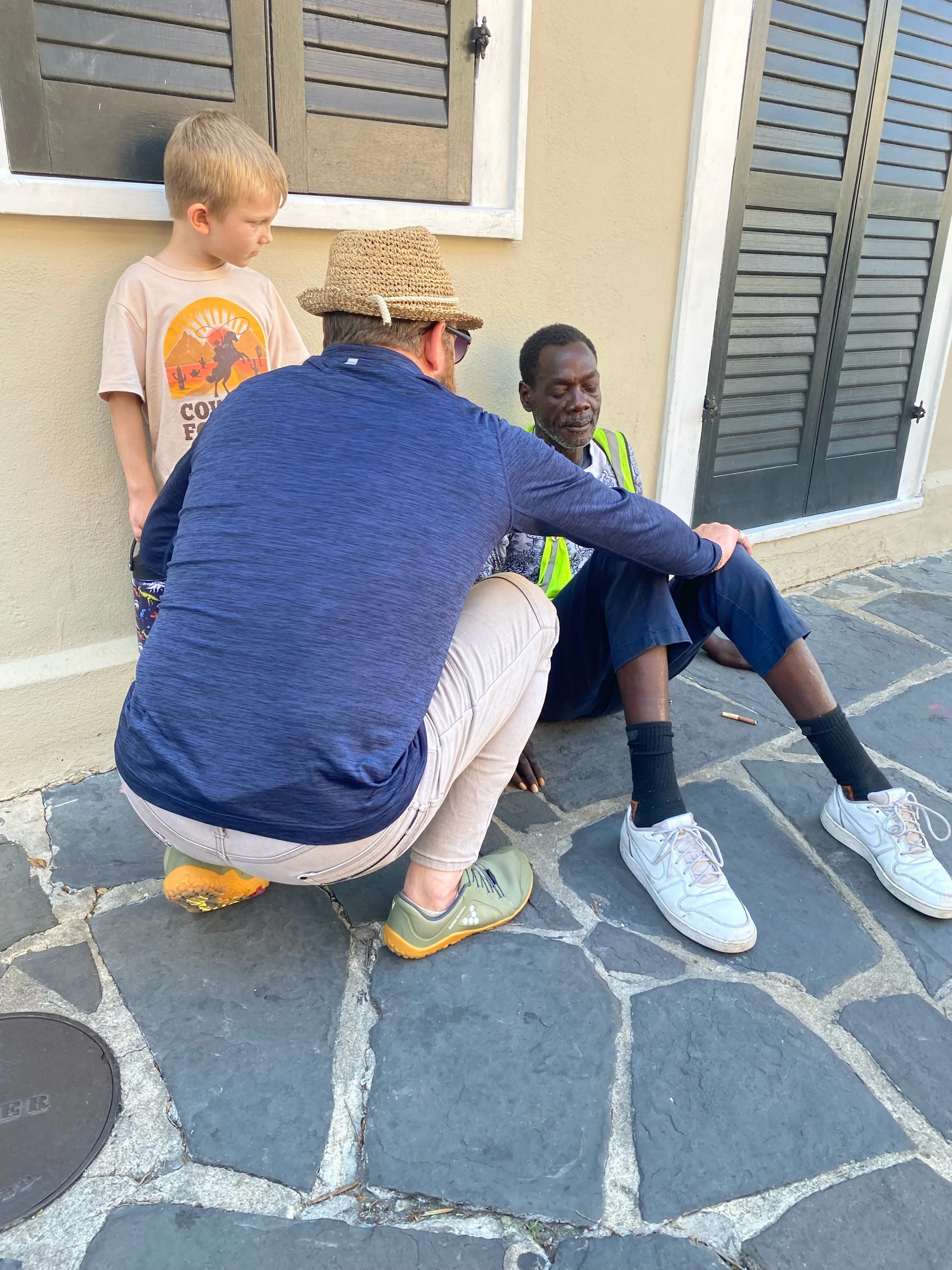 Two men and a young boy sitting and crouching on a stone sidewalk against a wall with black shutters. One man is sitting, wearing a reflective vest, while the other man prays and a boy observe.
