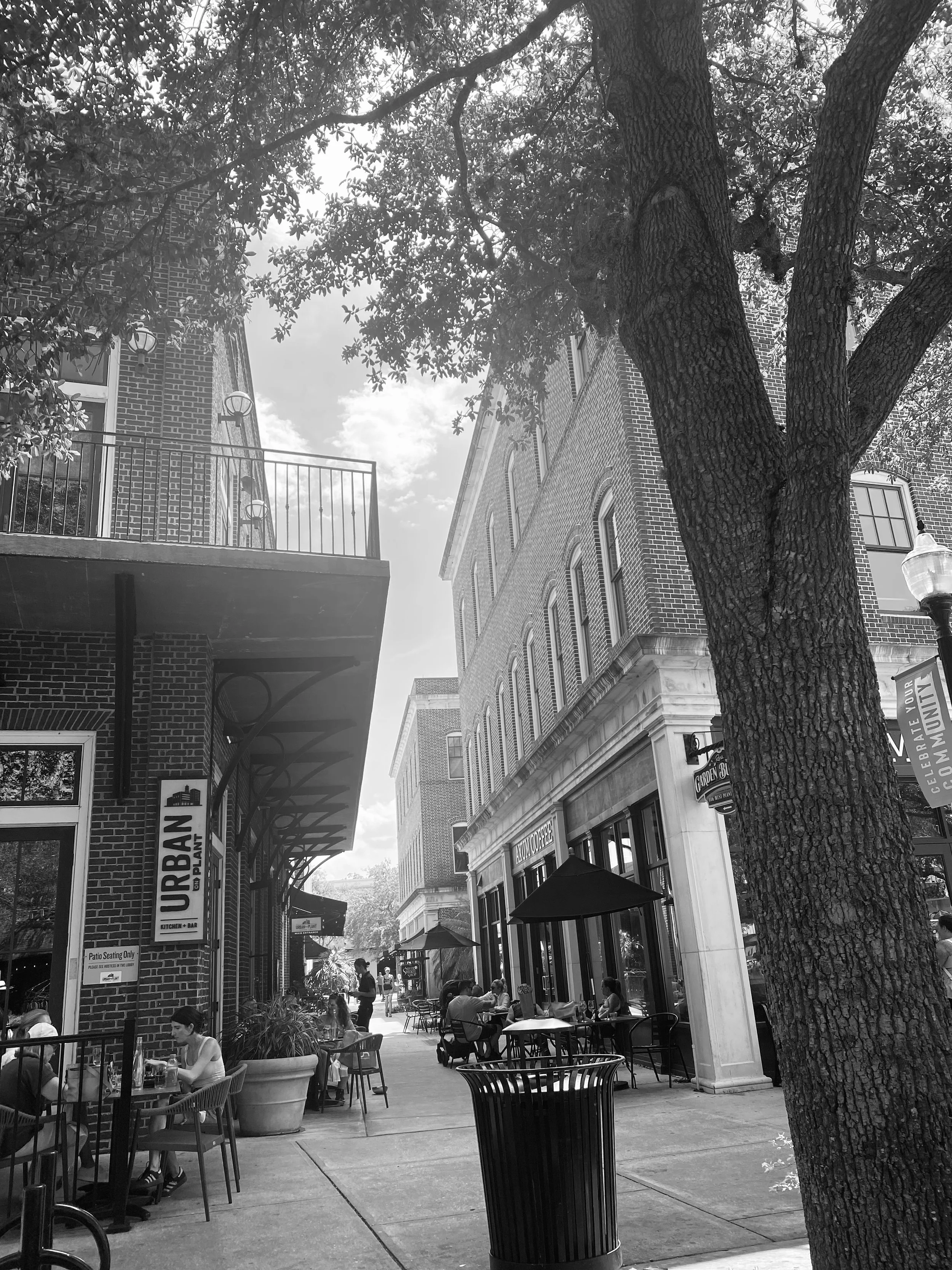 An outdoor city sidewalk restaurant with tables and chairs, some people dining, a large tree in the foreground, brick buildings, umbrellas, and a streetlamp.