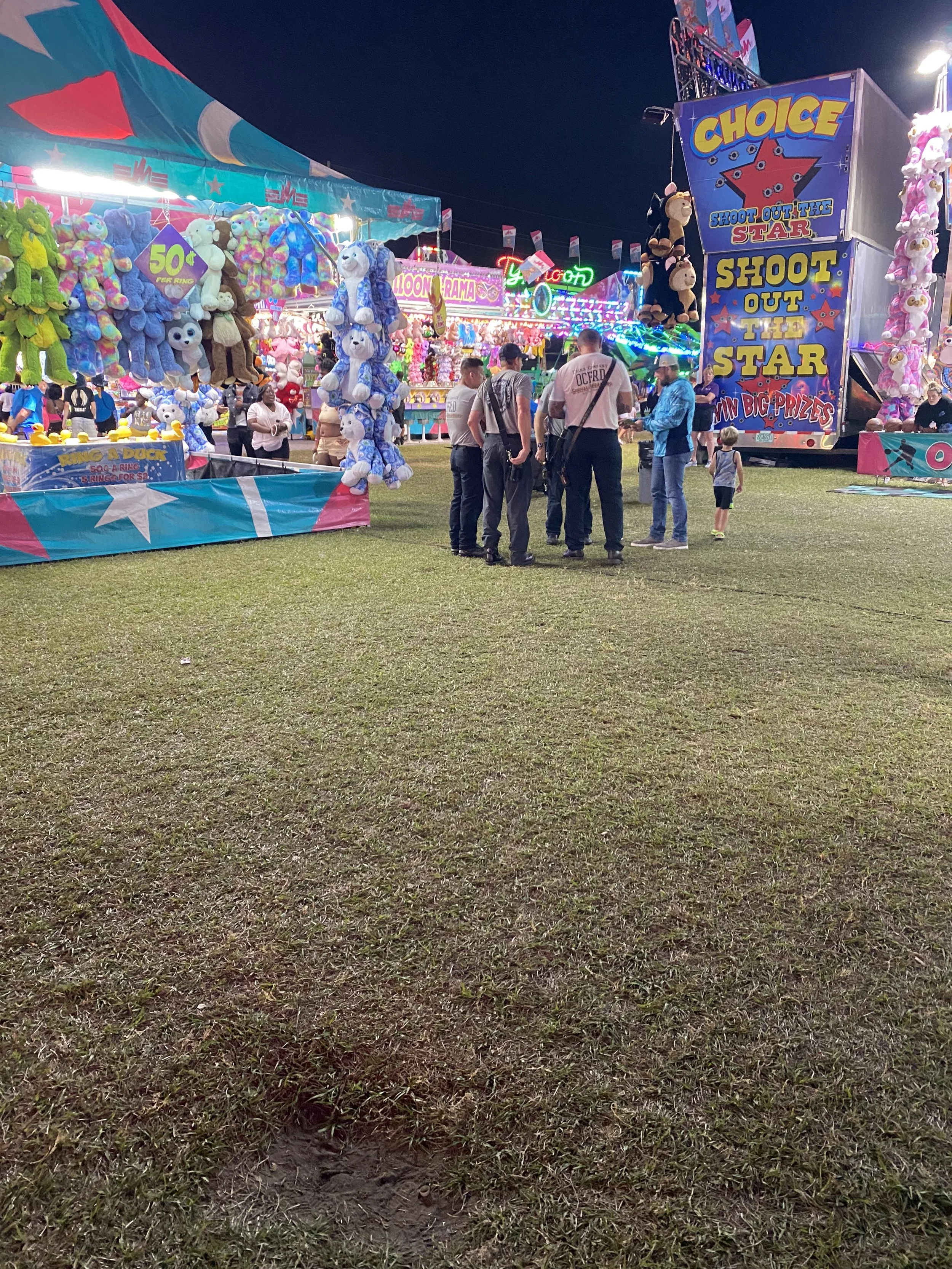 A night scene at a carnival with various game booths, bright lights, and a group of people standing and praying in front of a large shooting game. Colorful stuffed animals and prizes are displayed at the booths.
