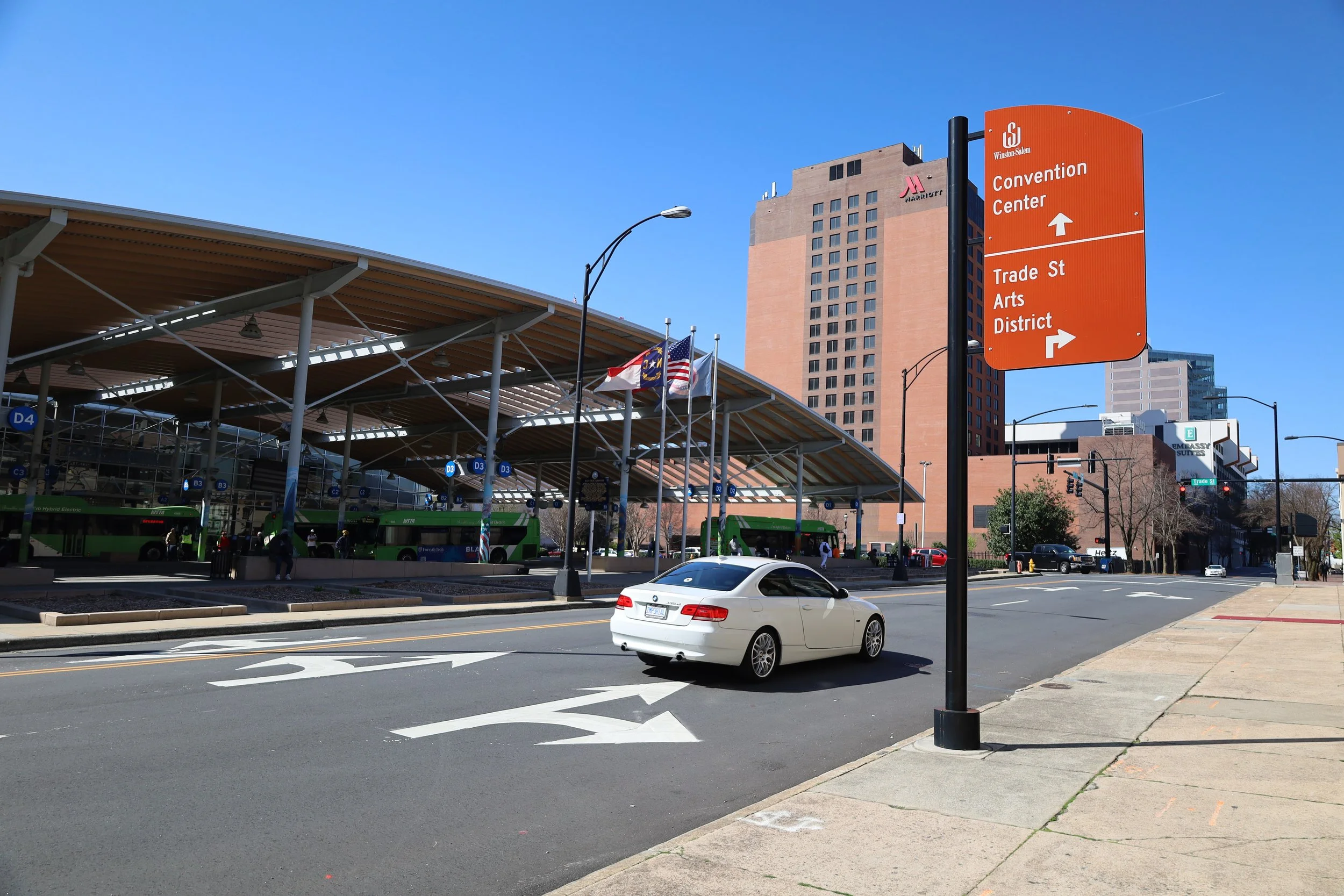 City street scene with a modern bus terminal, green buses, a white car, and tall buildings in the background. A bright orange directional sign indicates paths to the convention center and arts district.