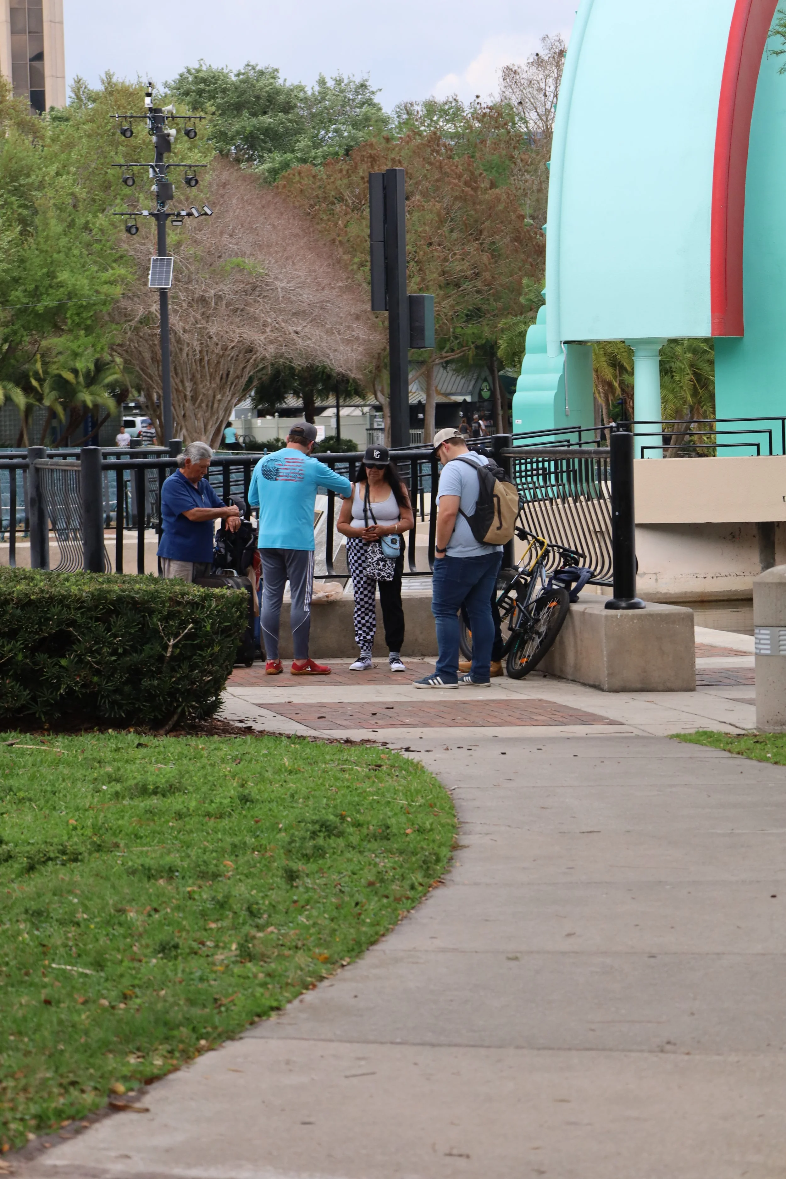 Group of five people standing and praying in a park near a sidewalk, with some bikes and trees in the background.