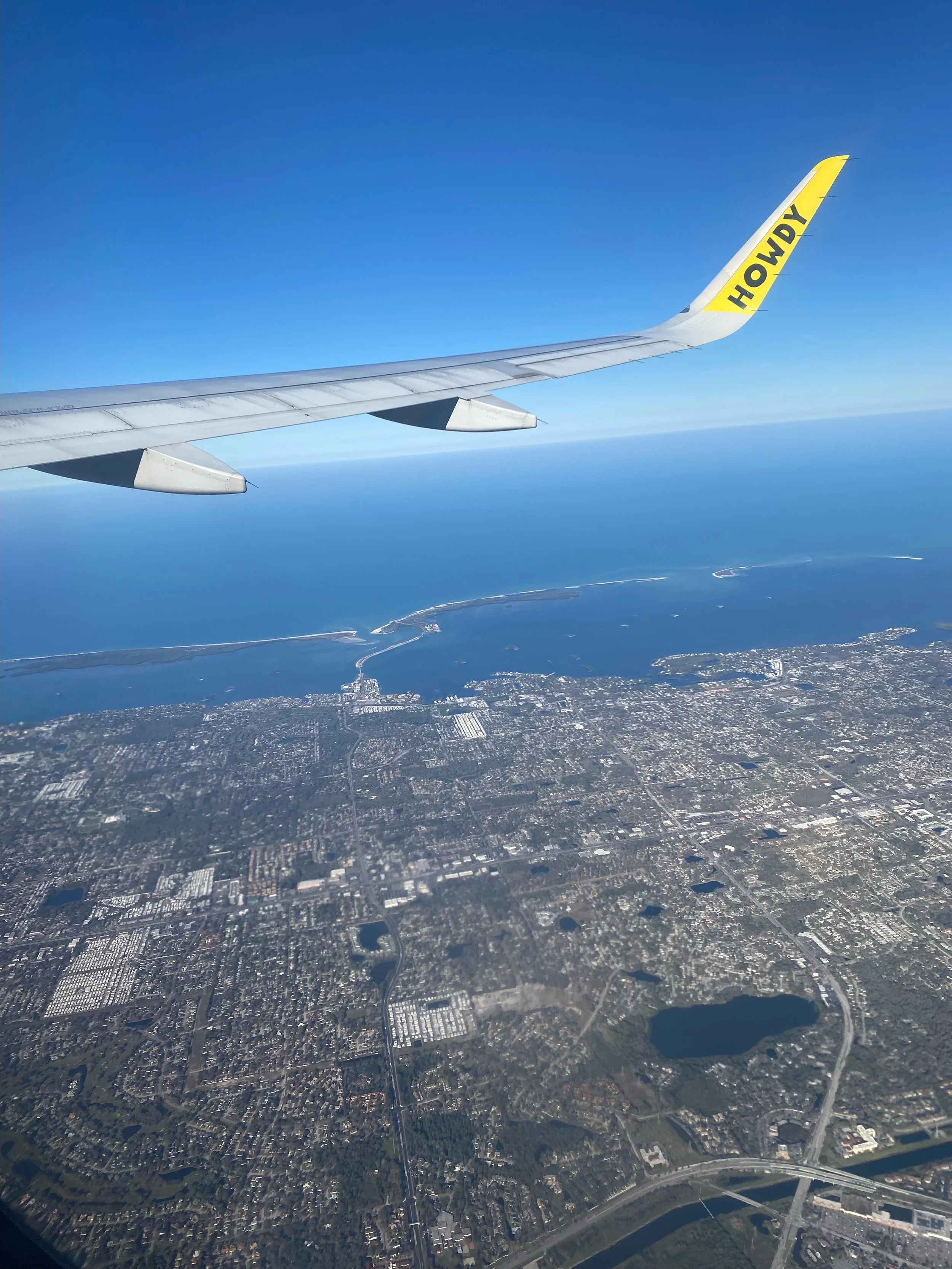 View from airplane window showing wing with the word 'HOWDY' on the wingtip, over a city with water bodies and islands below.