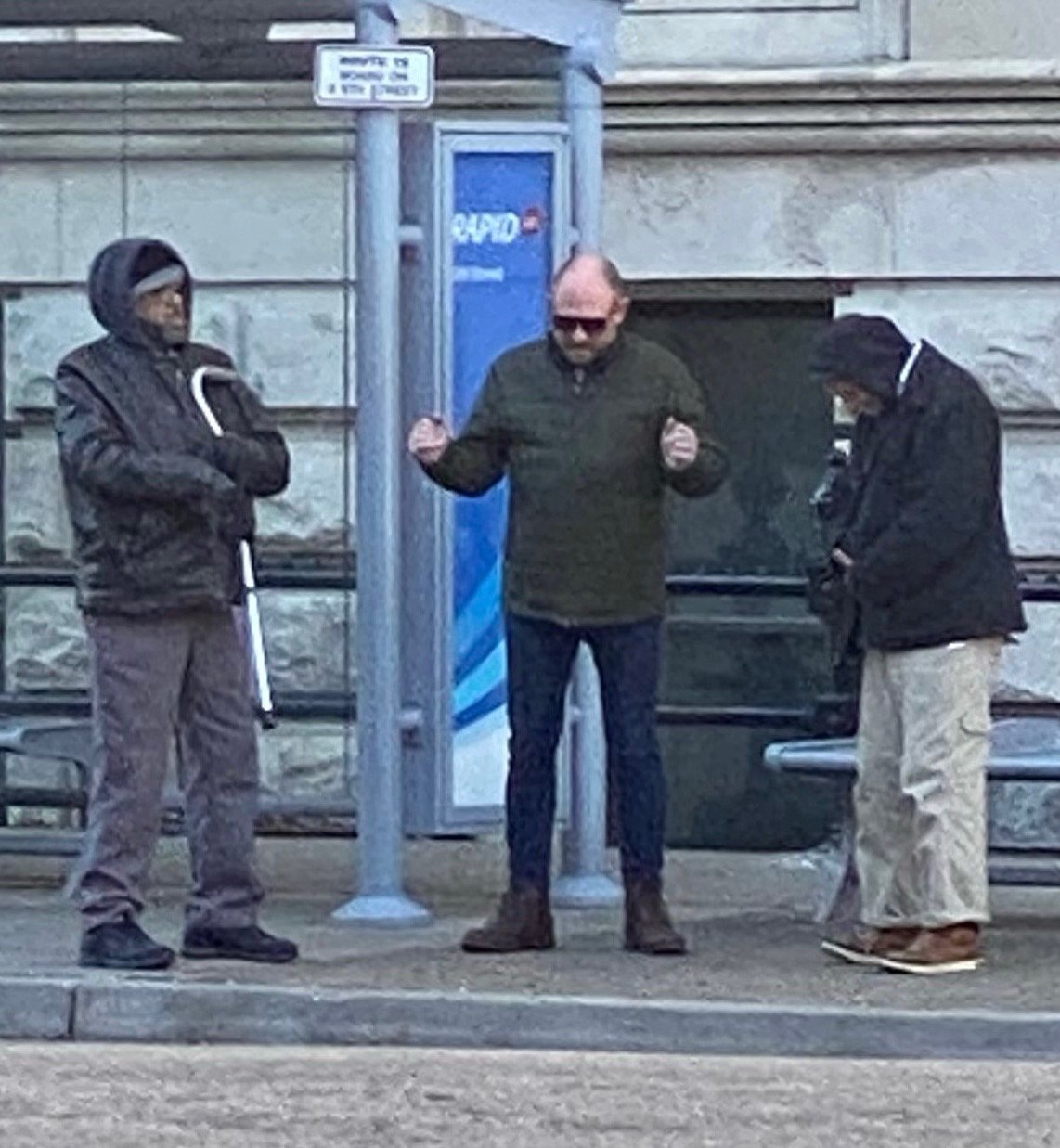 Three men standing at a bus stop on a city sidewalk, one wearing a hooded jacket, another in a green jacket with sunglasses, and the third with a black jacket, near a blue payphone.