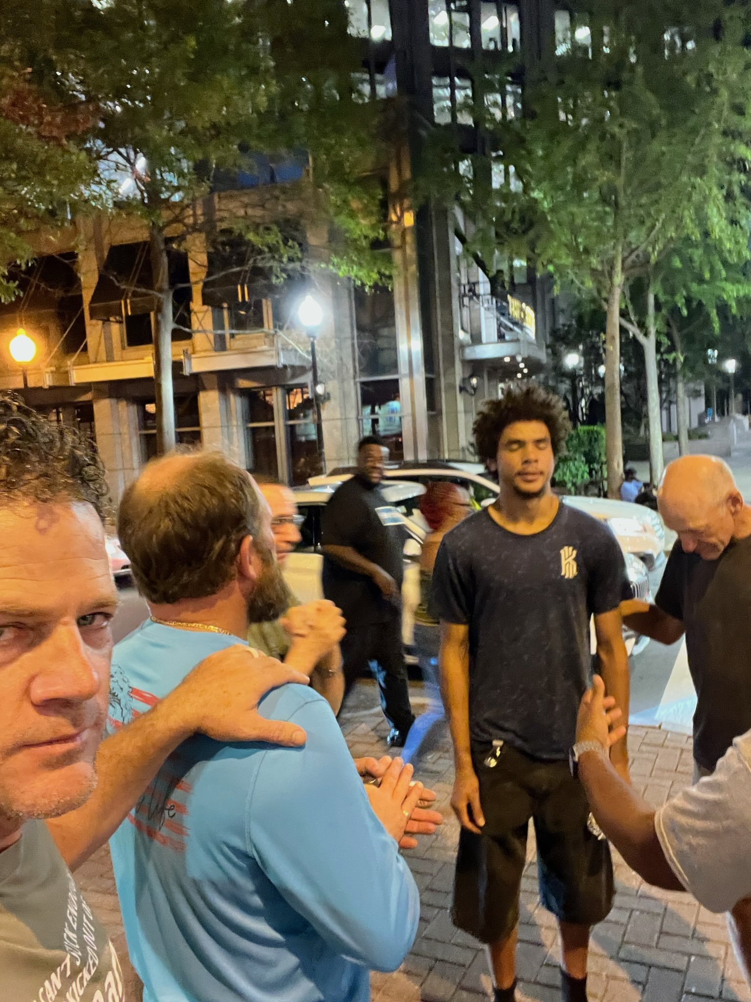 A group of diverse people standing on a city sidewalk at night engaging in prayer, with trees, buildings, and parked cars in the background.