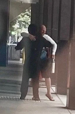 Two people embracing, one with their back to the camera, standing near a glass door entrance outside a building.