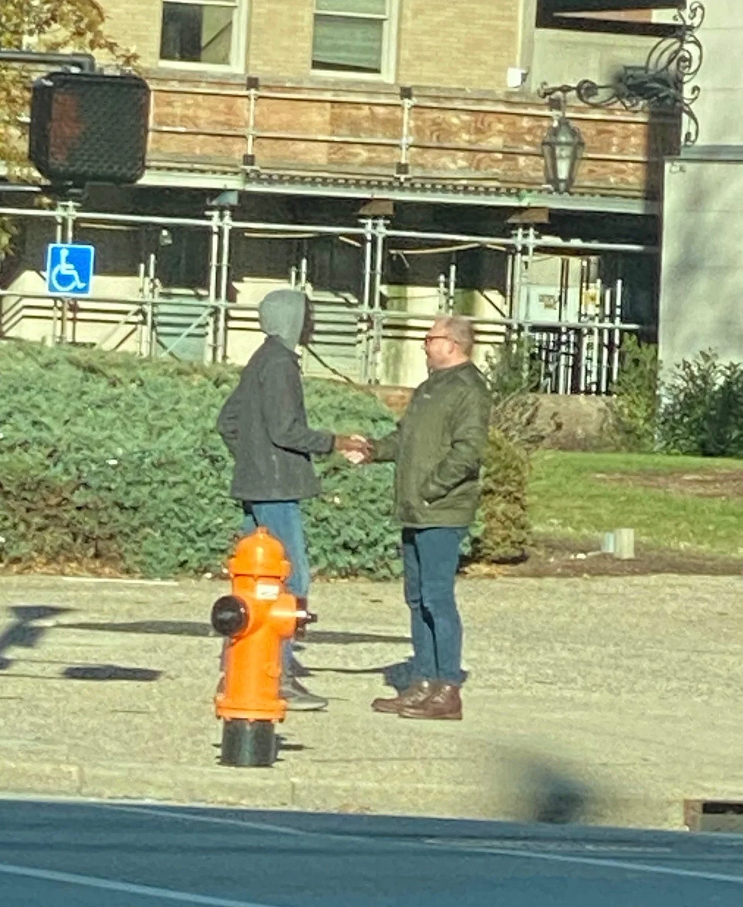Two men shaking hands on sidewalk near an orange fire hydrant. One man is wearing a gray hoodie and jeans, the other is wearing glasses and a green jacket.
