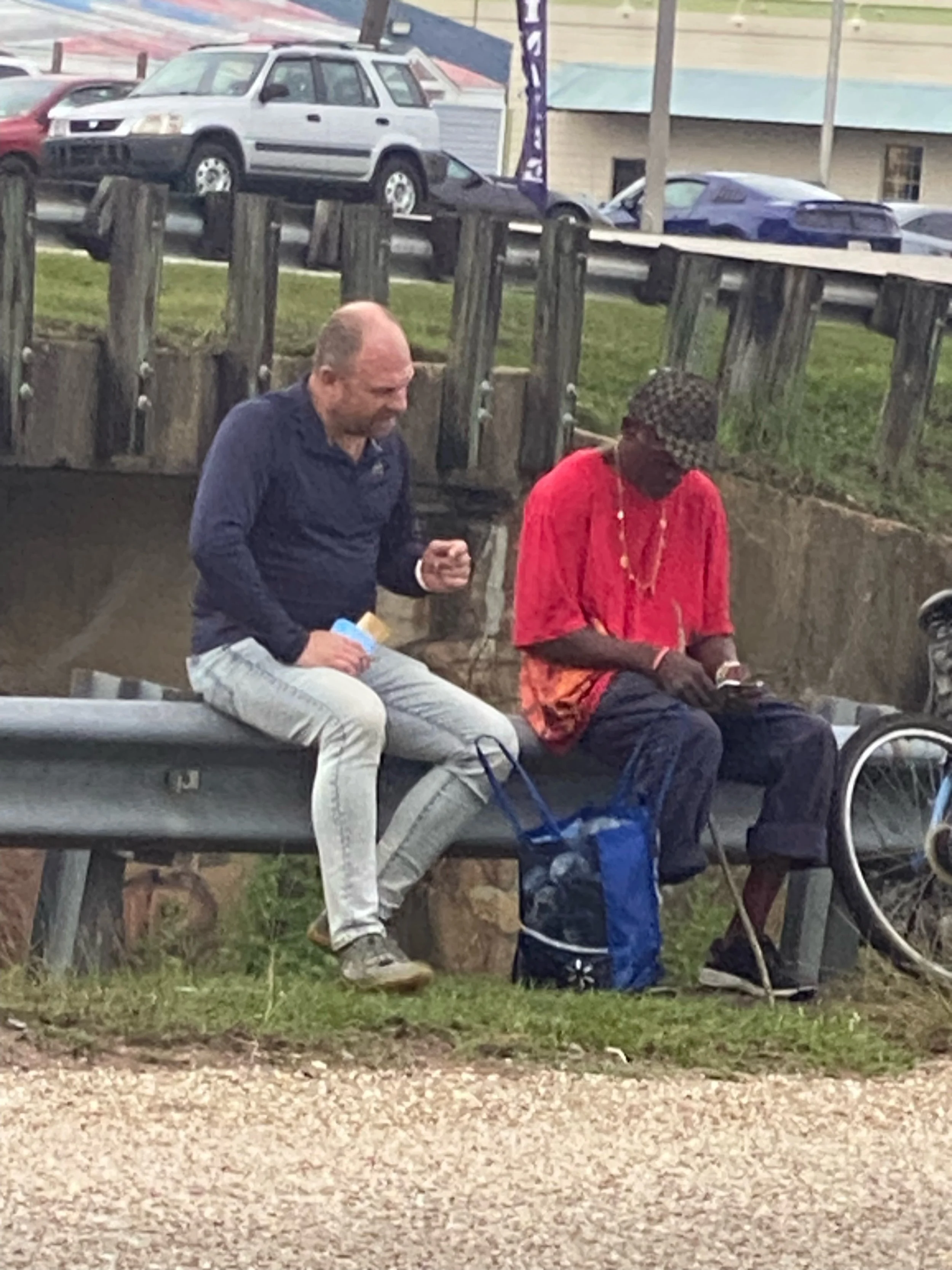 Two men sitting on a park bench talking and praying. Cars are parked in the background.