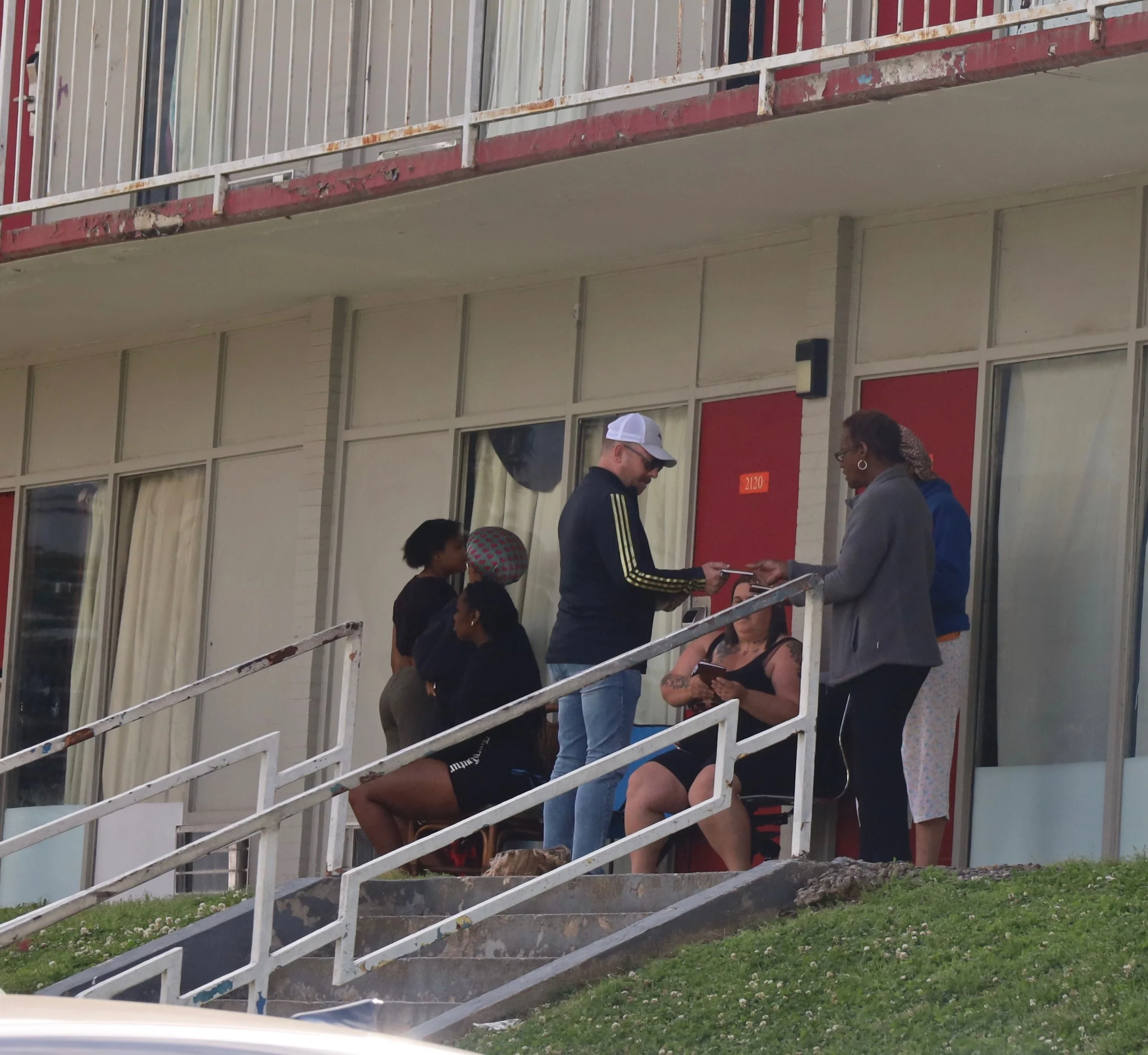 A group of six people gathered on the stairs outside an apartment building, with some sitting and some standing, engaging in prayer, while others wait nearby.