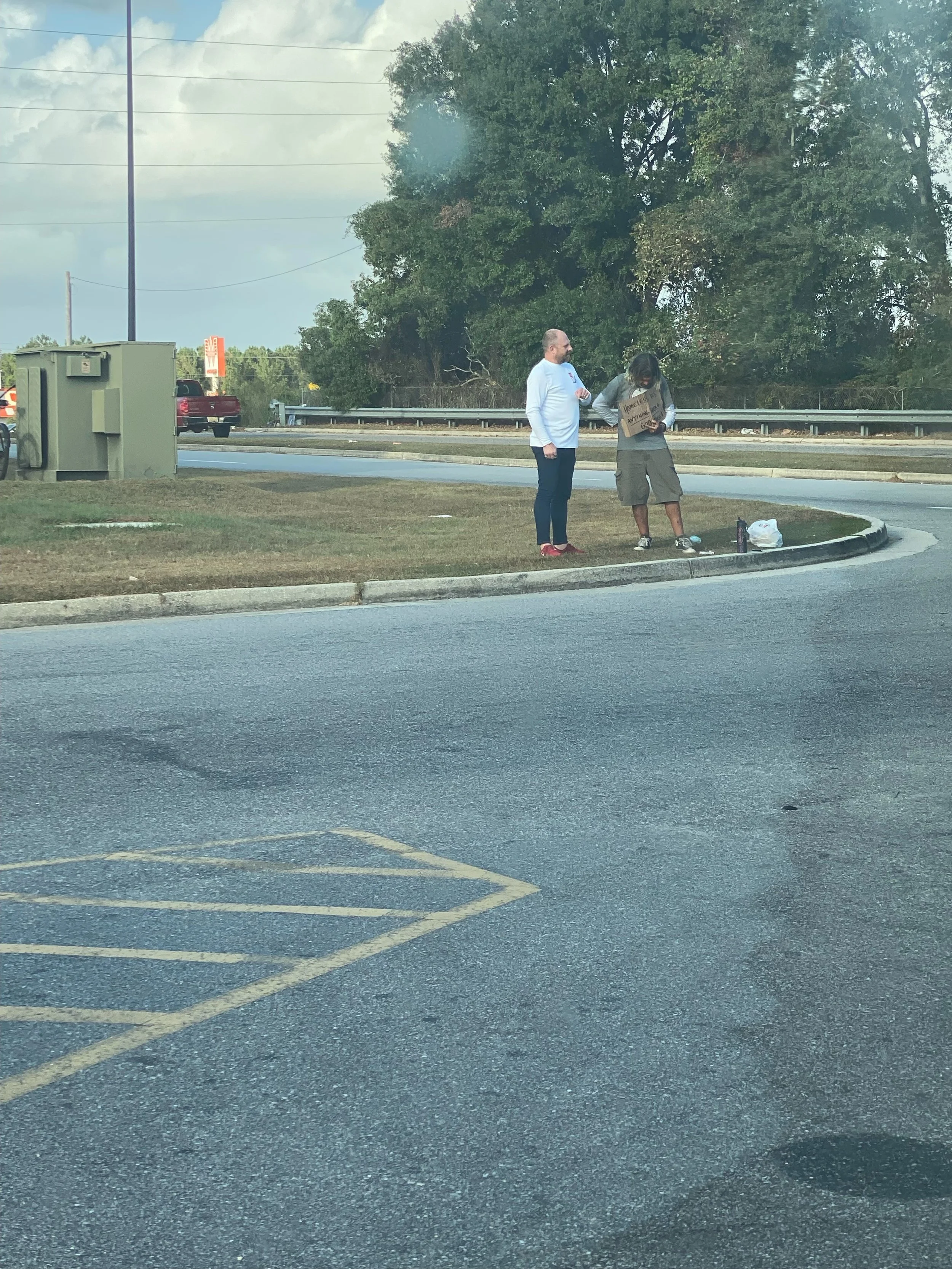 Two people standing on a sidewalk near a road, one holding a cardboard sign, with a tree in the background and some belongings on the ground.