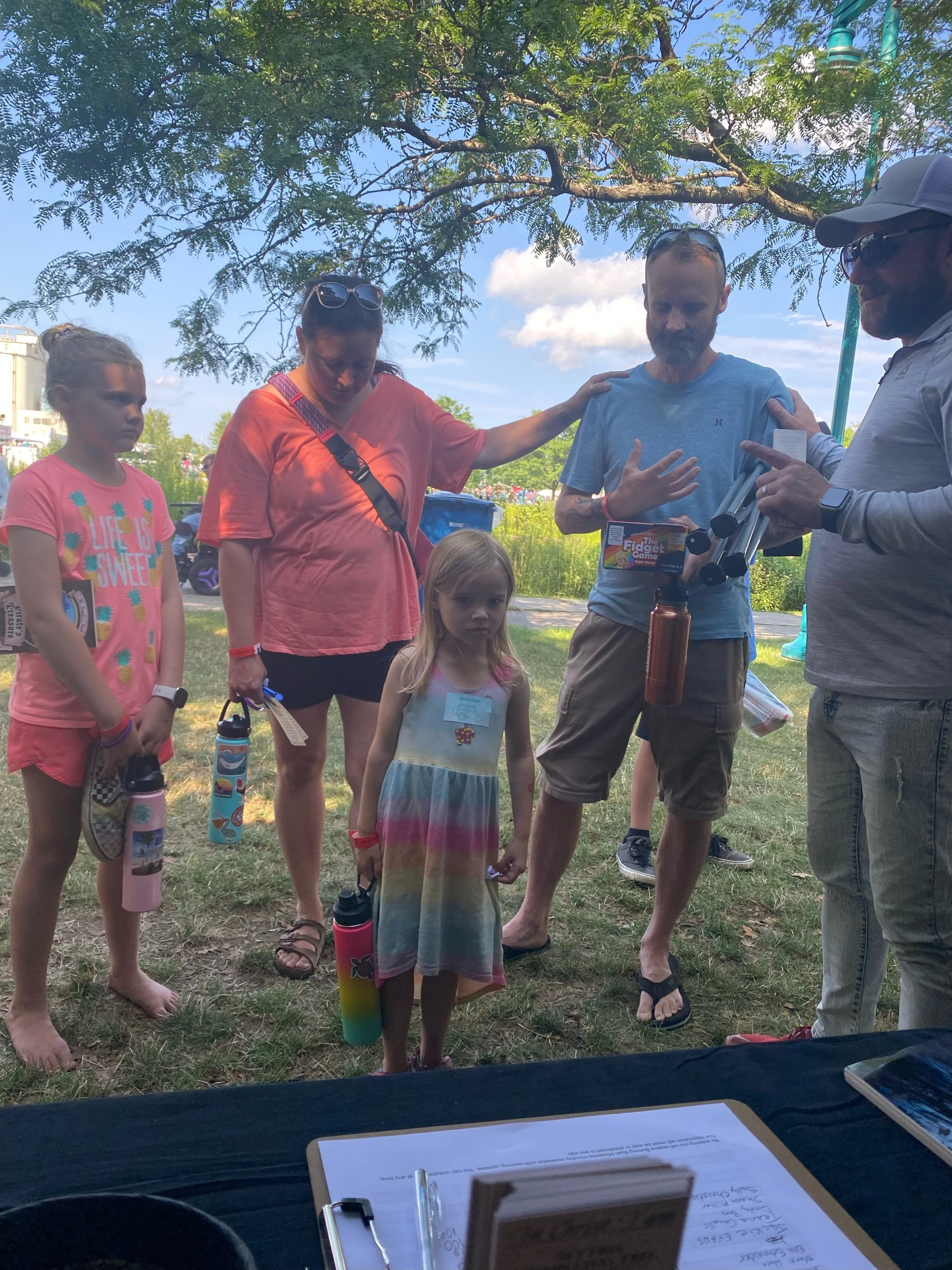 A group of children and adults outdoors under a tree, with some holding water bottles, engaging in prayer during a sunny day.
