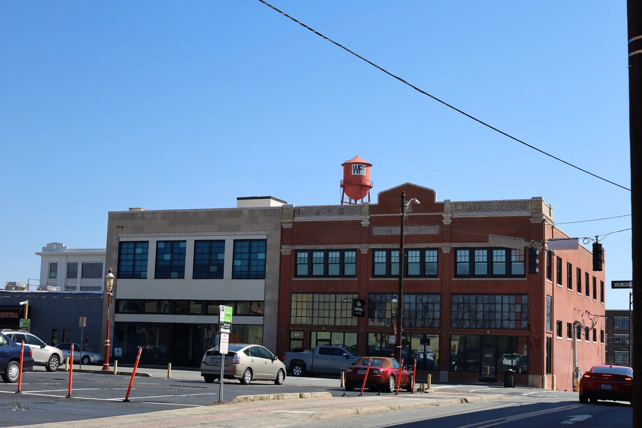 A street view showing two brick buildings with storefronts and large windows, parked cars, street lamps, and utility poles, with a clear blue sky in the background.