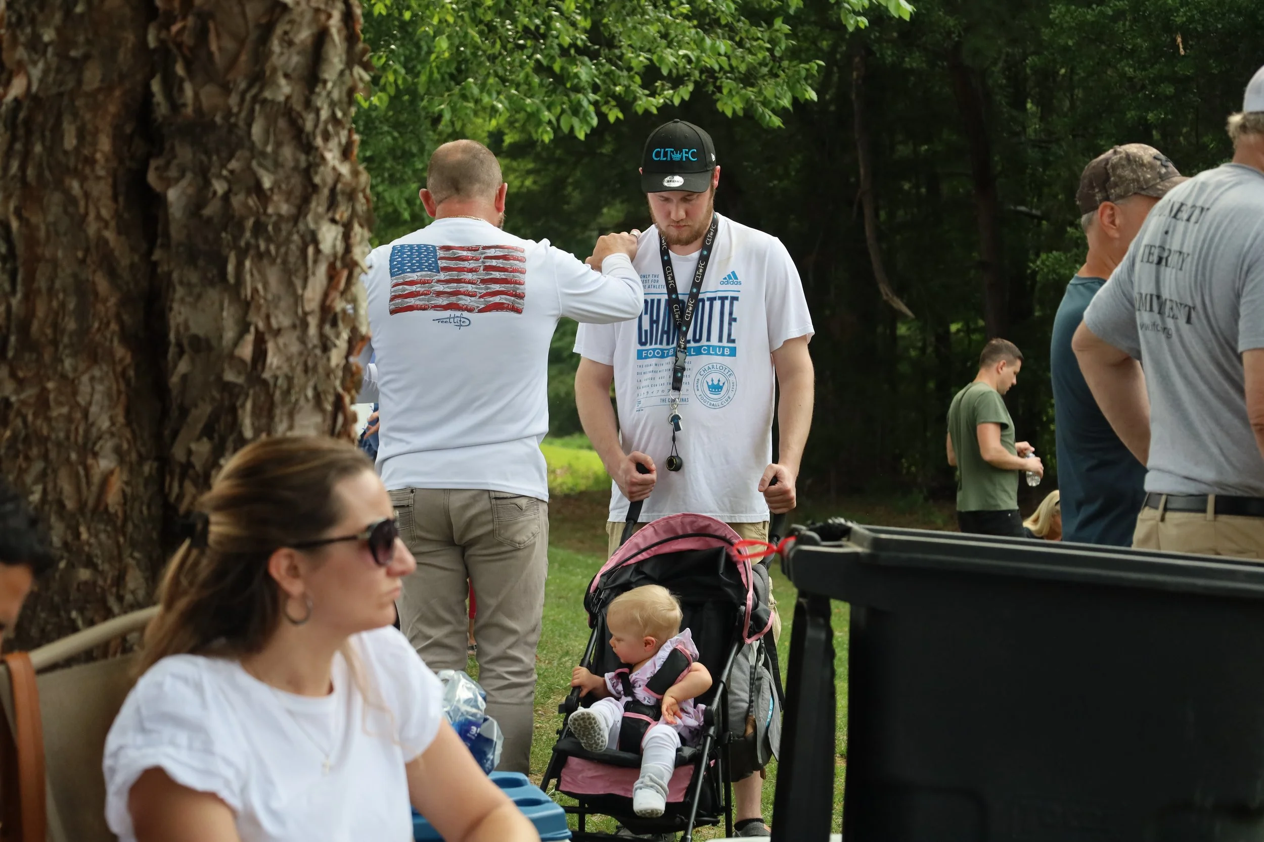 A group of people outdoors in a park, including a man in a Charlotte FC t-shirt, holding a stroller with a young girl inside and praying with a man. A woman wearing sunglasses sitting nearby, with other individuals are in the background near trees.