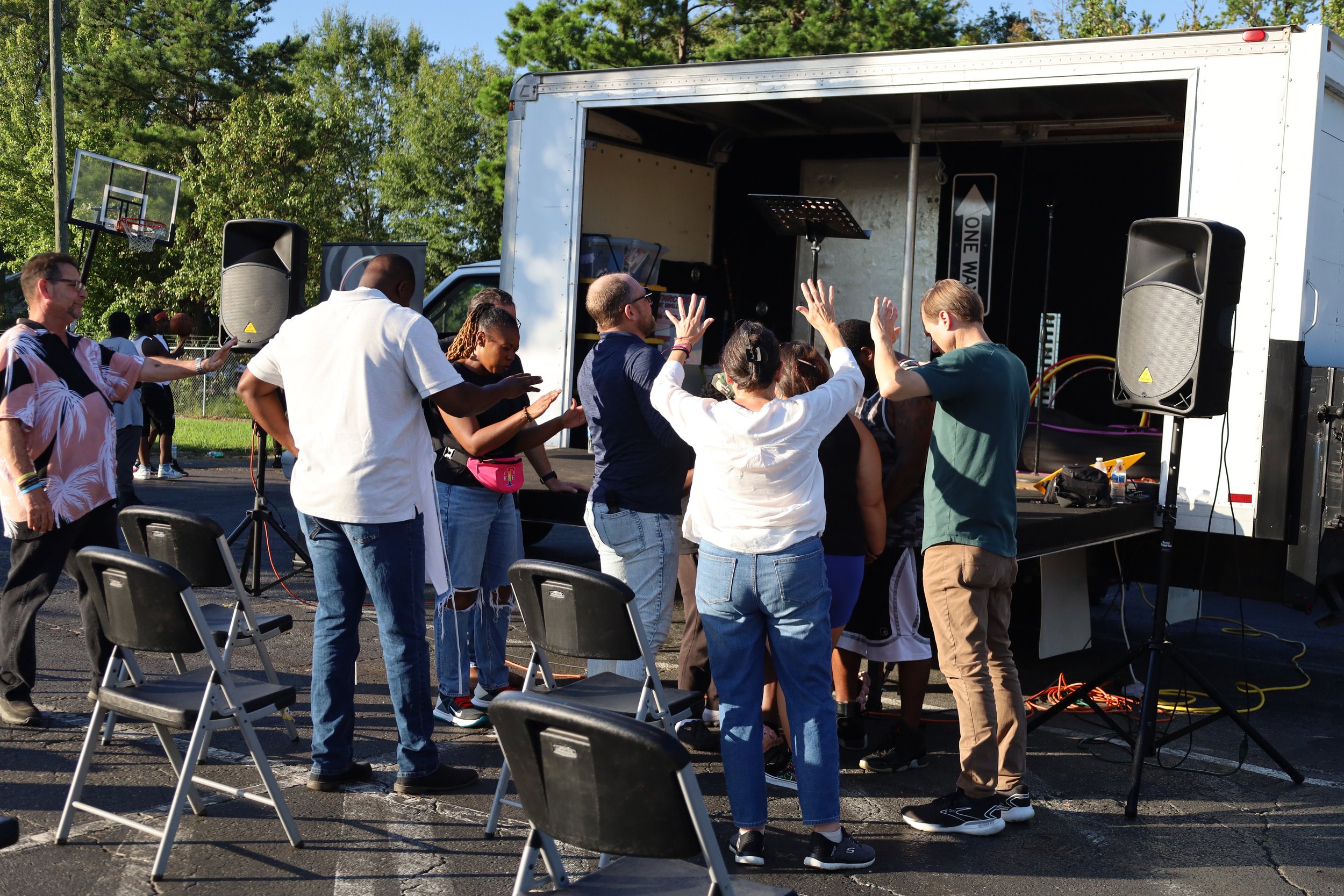 People gathered around a mobile stage with musical equipment, some with their hands raised praying, outdoors on a sunny day with green trees in the background.