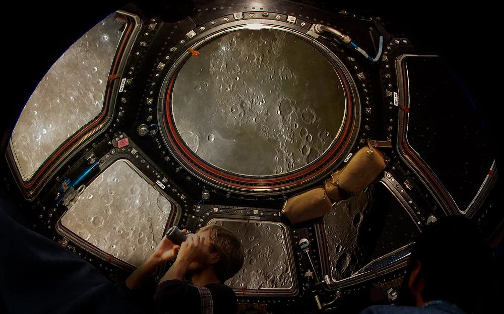Tourists float in a version of the ISS Cupola module with the surface of the Moon out the windows.