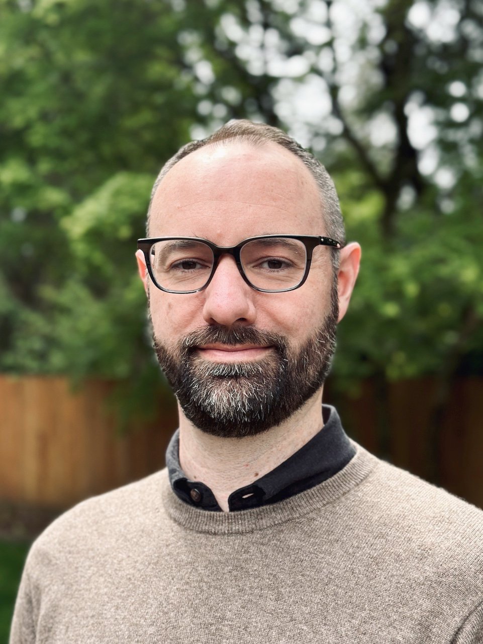 Portrait of a man with glasses and a beard, standing outdoors with trees and a wooden fence in the background.