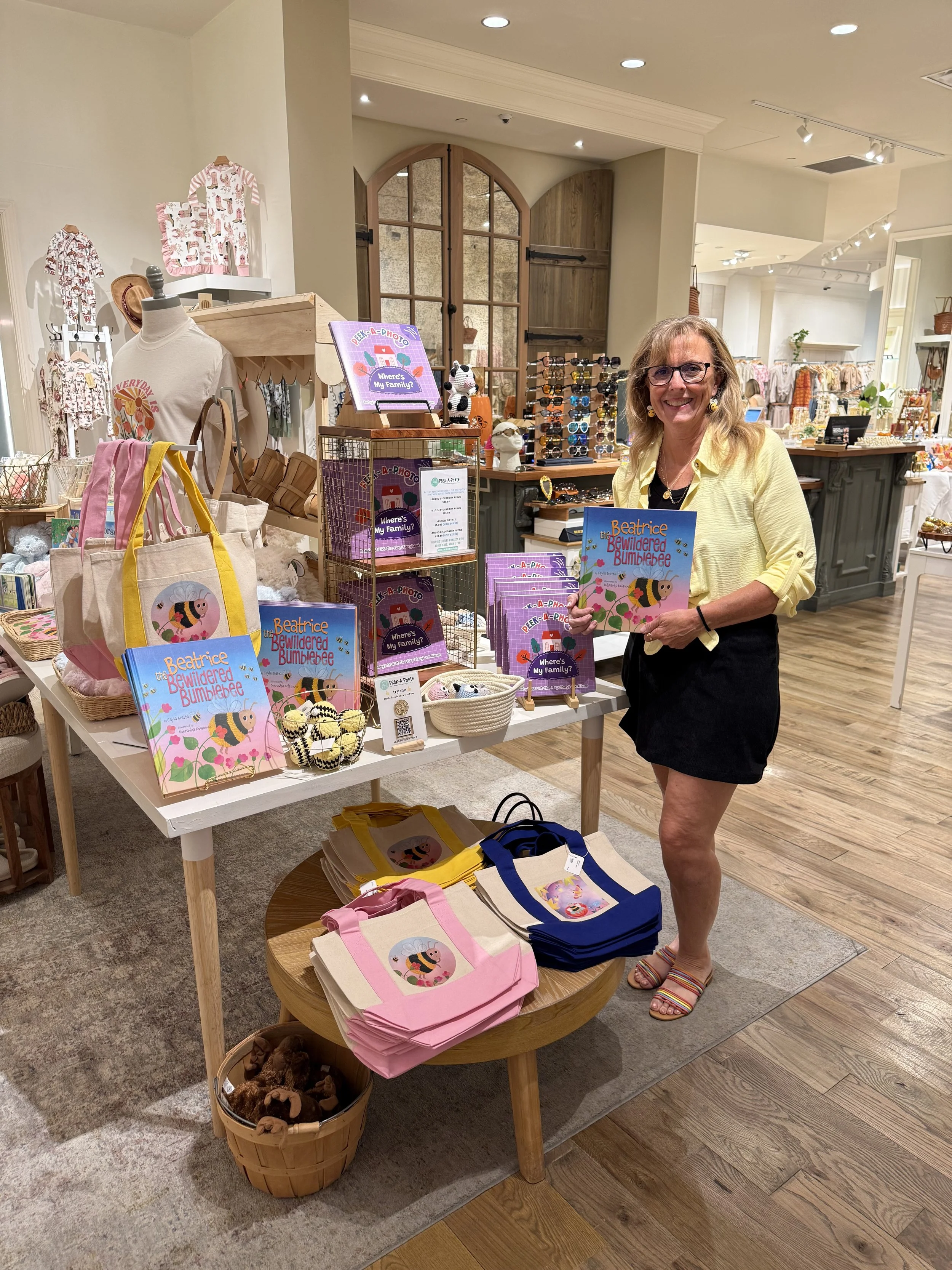 A woman smiling and holding a children's book titled 'The Bewildered Bumblebee' at a retail store display with various bags, books, and accessories related to a bee theme.