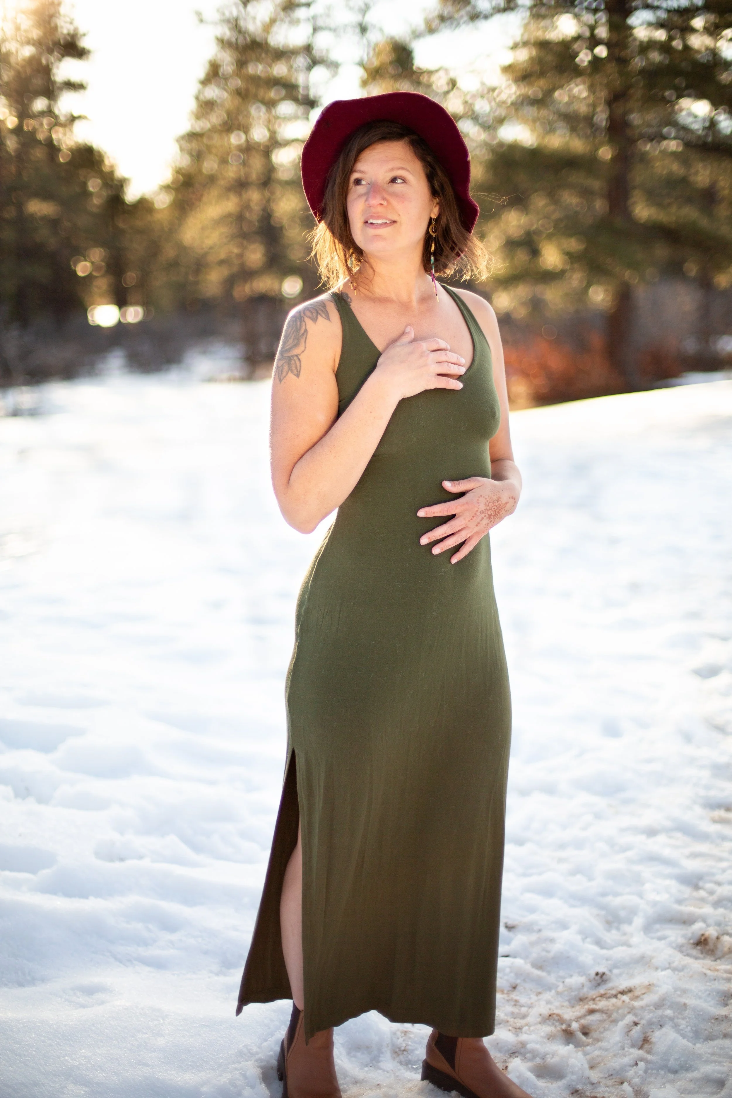 Woman in a burgundy hat and olive green dress standing in snowy woods.