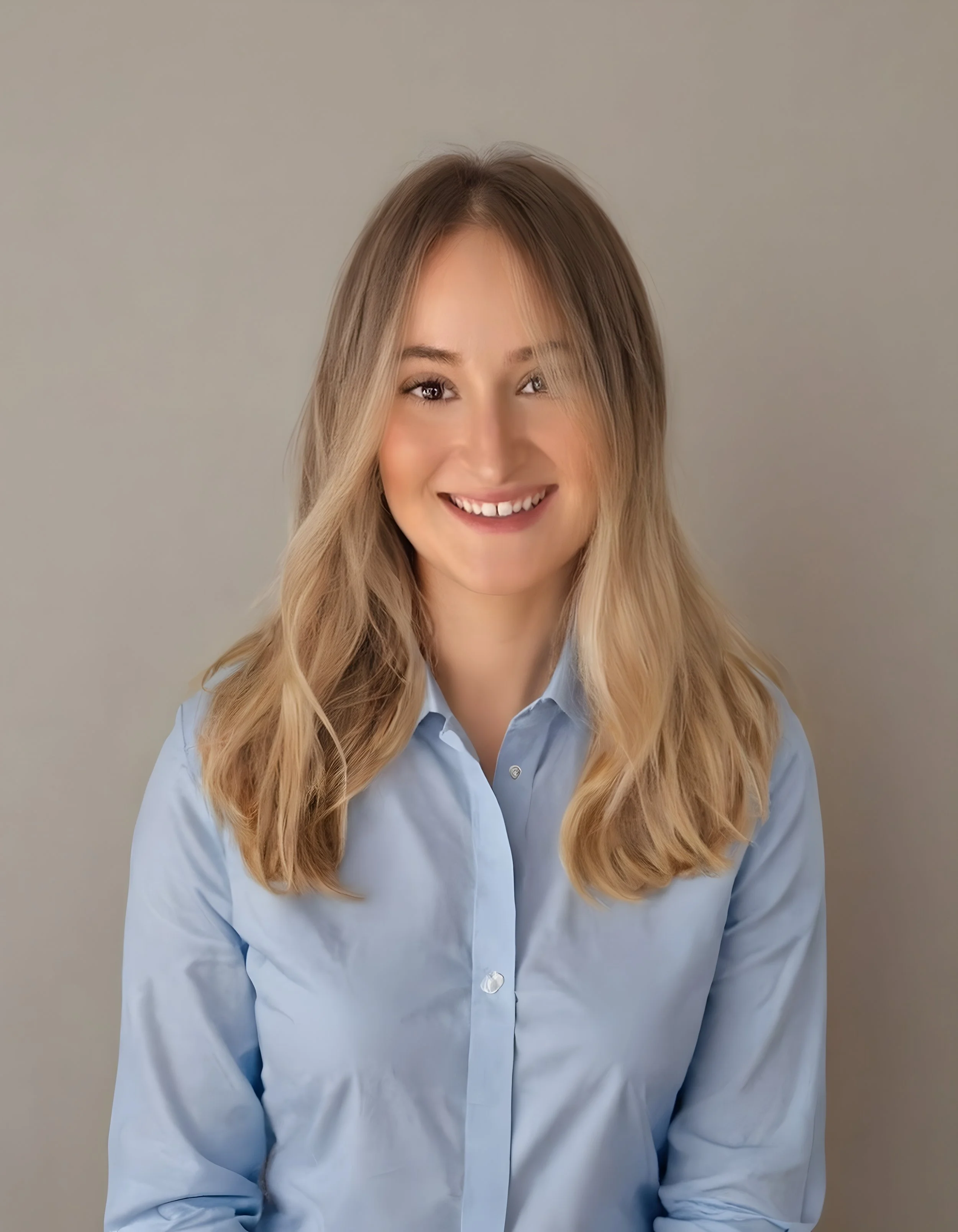 A smiling young woman with long, wavy blonde hair wearing a light blue button-up shirt, standing against a plain neutral background.