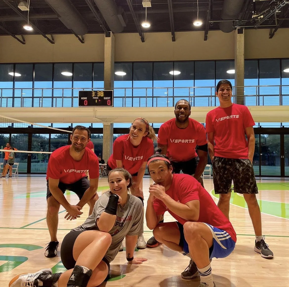 Six people pose and smile on an indoor volleyball court; five wear matching red shirts, one in gray kneels in front.