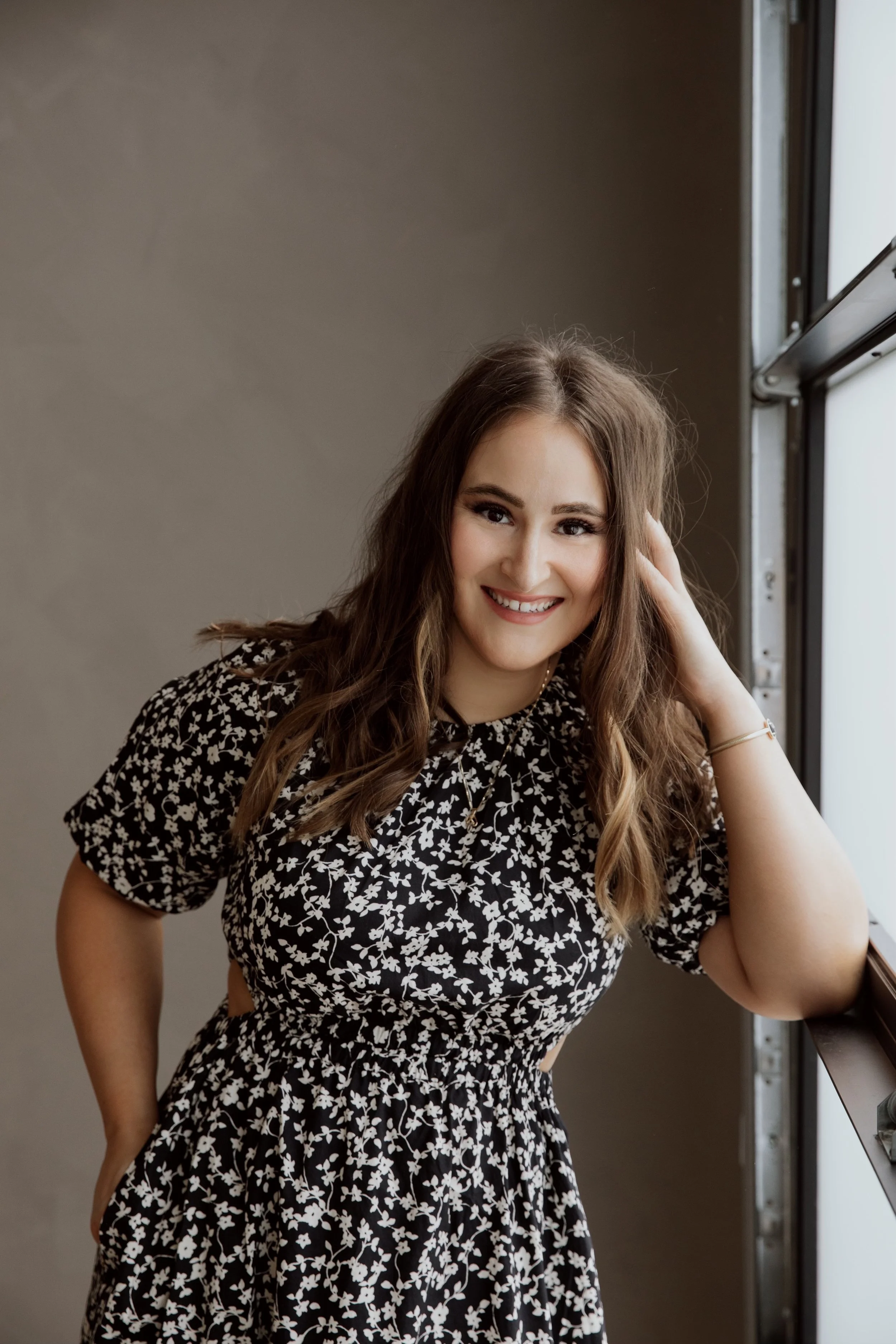 A young woman with long brown hair, smiling, posing near a window, wearing a black and white floral dress.