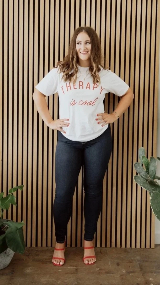 Founder of Own The Moment, Stephanie Lindsey standing in front of a wooden textured background. She is smiling with her hands on her hips while wearing a "therapy is cool" t-shirt.