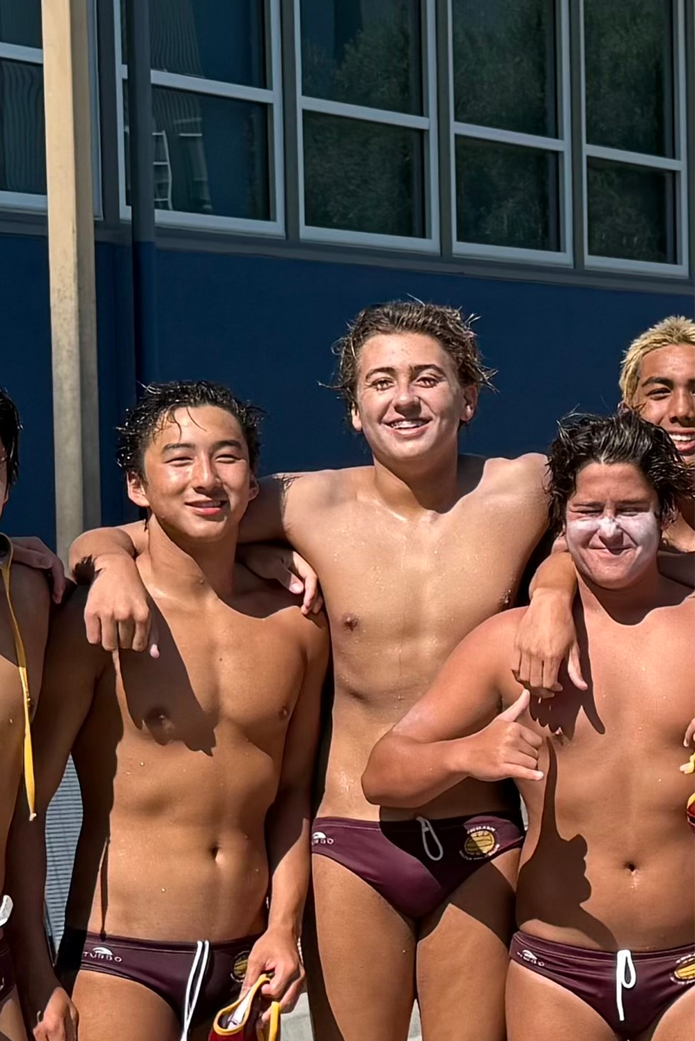 Group of young male water polo players shirtless, smiling, with their arms around each other, outdoors near a building with large windows.