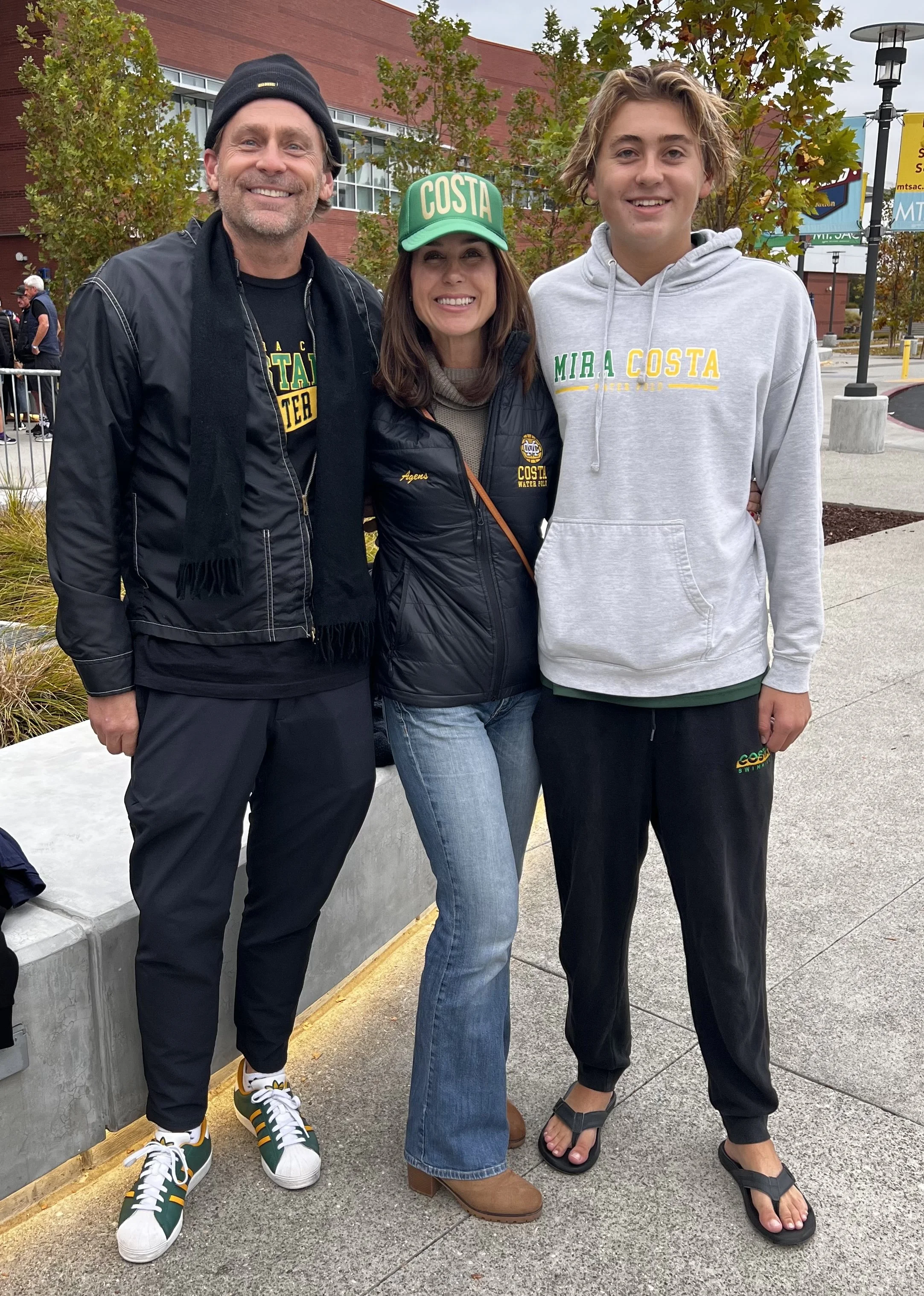 Three people standing outdoors, smiling for a photo. The group includes a man in a black beanie and athletic wear, a woman in a black jacket and green cap, and a teenage boy in a gray hoodie and black pants. They are in front of a building with red b