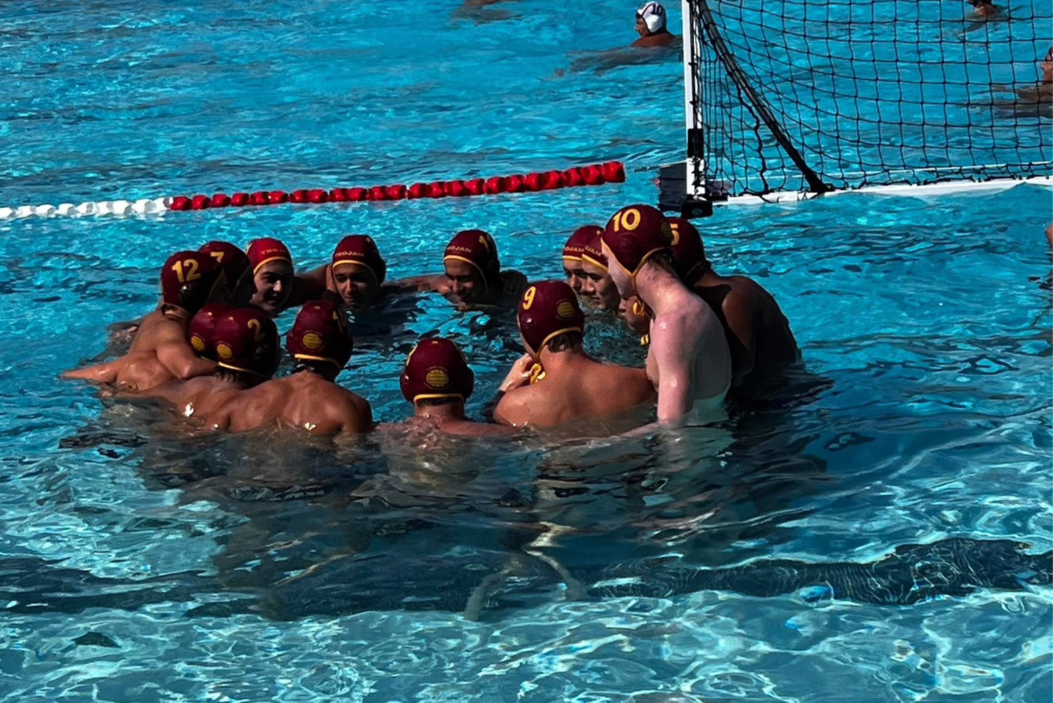 Water polo team huddled together in a pool, possibly during a timeout or team talk, wearing matching maroon caps with yellow markings and numbers, near a goal.