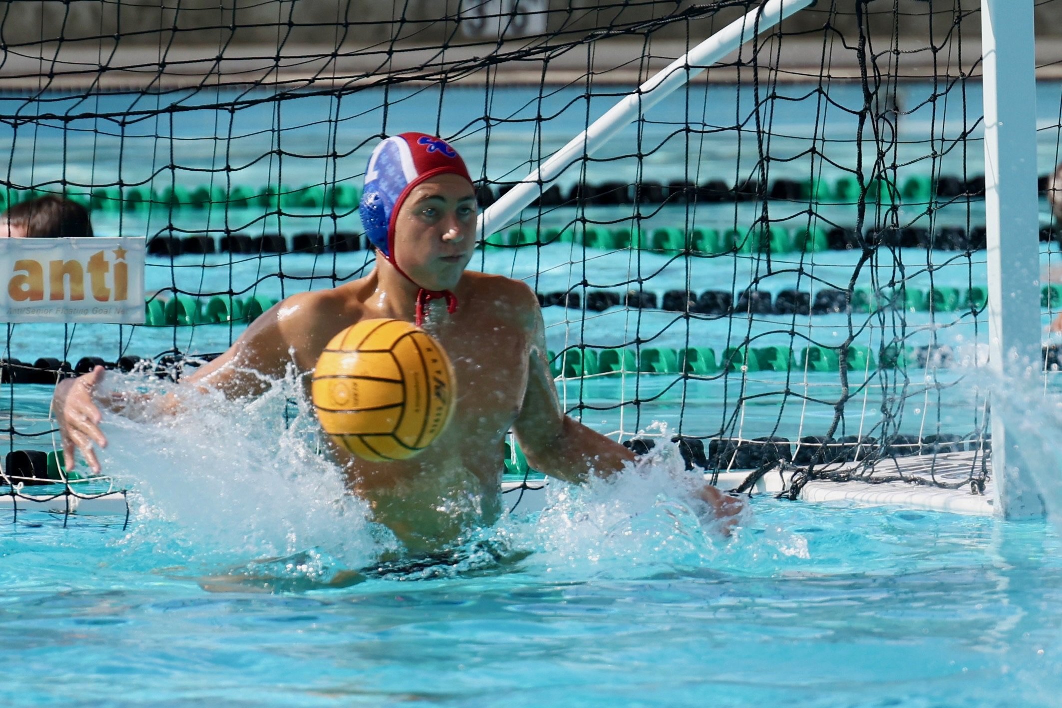 Water polo goalkeeper with red, white, and blue cap in a swimming pool, about to block a yellow water polo ball.