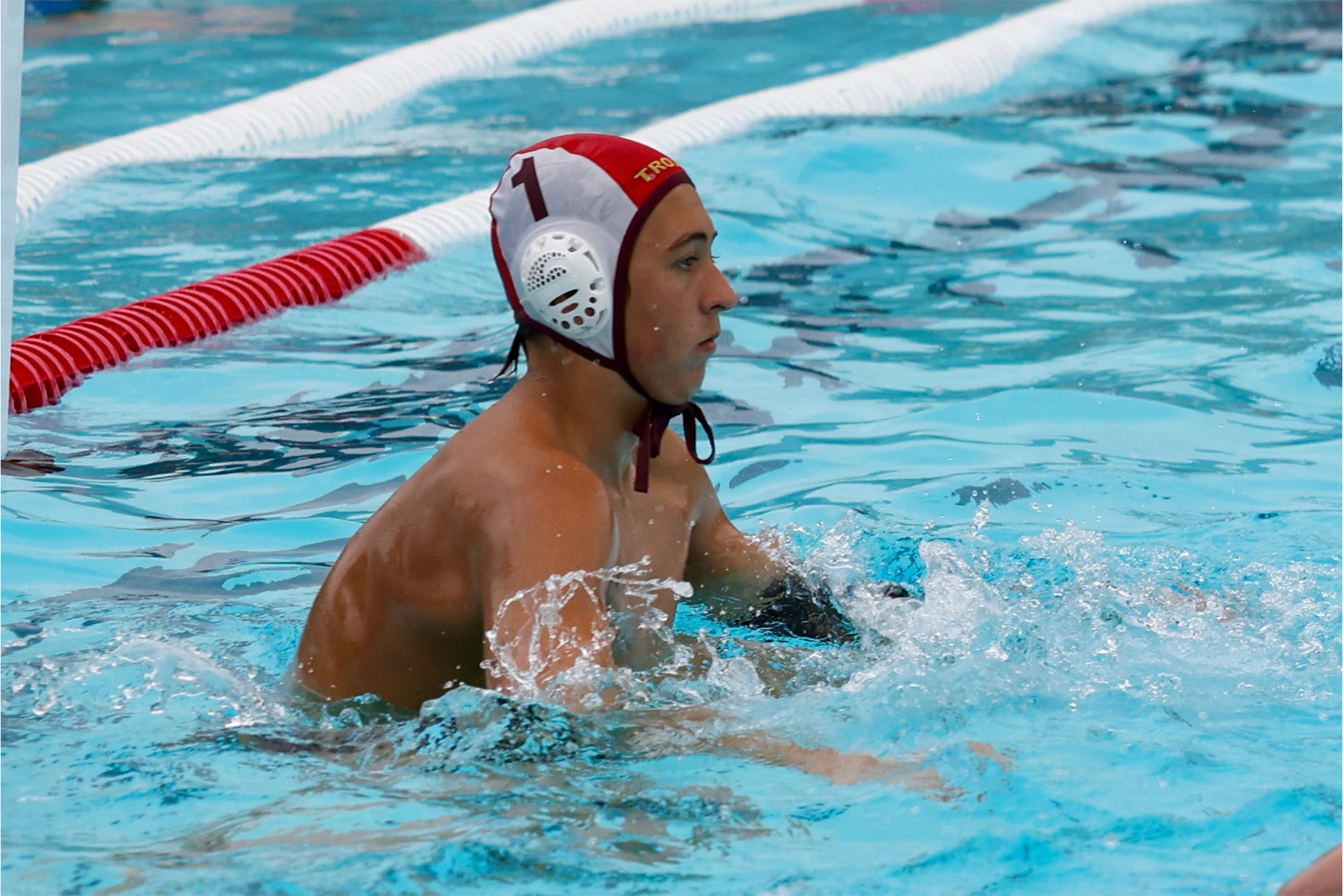 A water polo player with a white and red cap, with the number 1, in a swimming pool, appearing focused.
