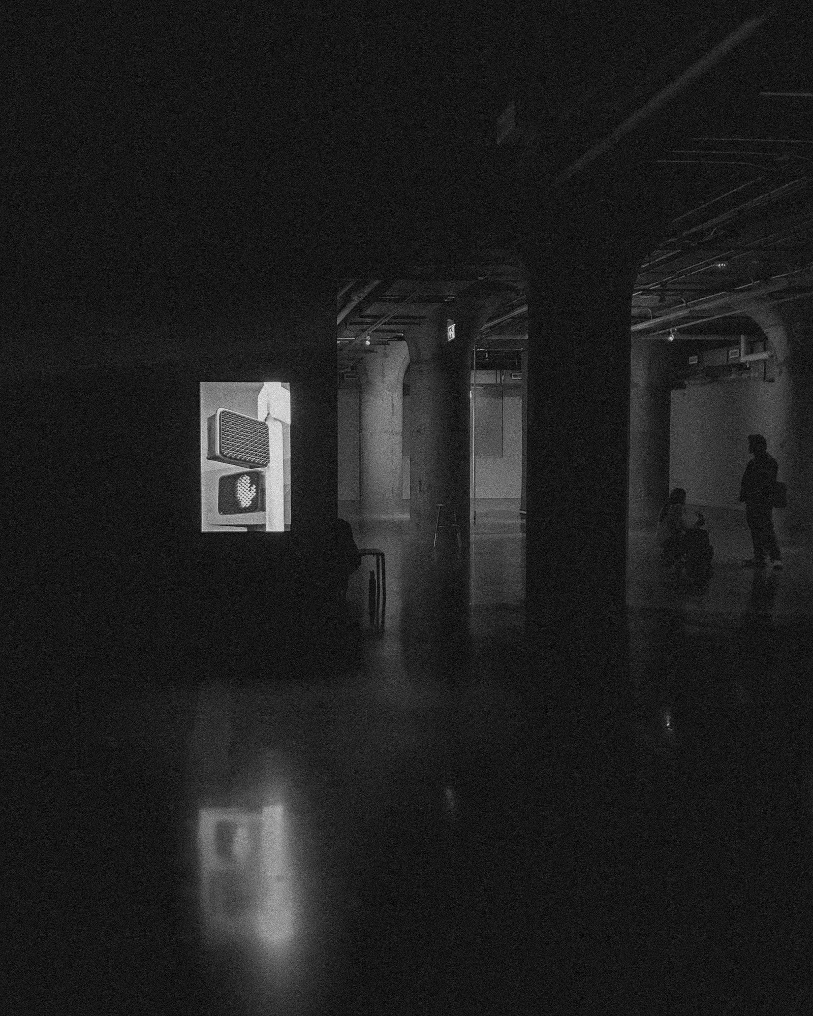 Black and white photograph of a dim gallery interior with artwork, reflections, and silhouetted visitors.
