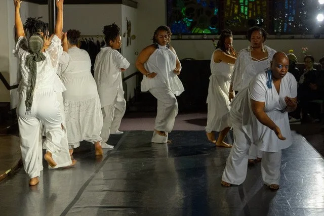Group of women dressed in white performing a dance on a dark stage indoors.