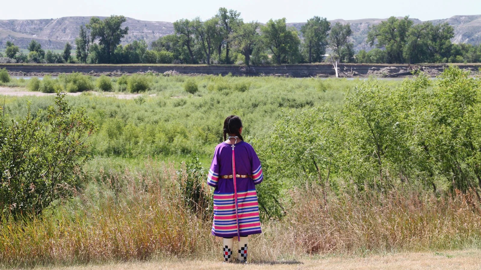 A person dressed in traditional Native American clothing standing outdoors, facing away, with green bushes, trees, and mountains in the background.