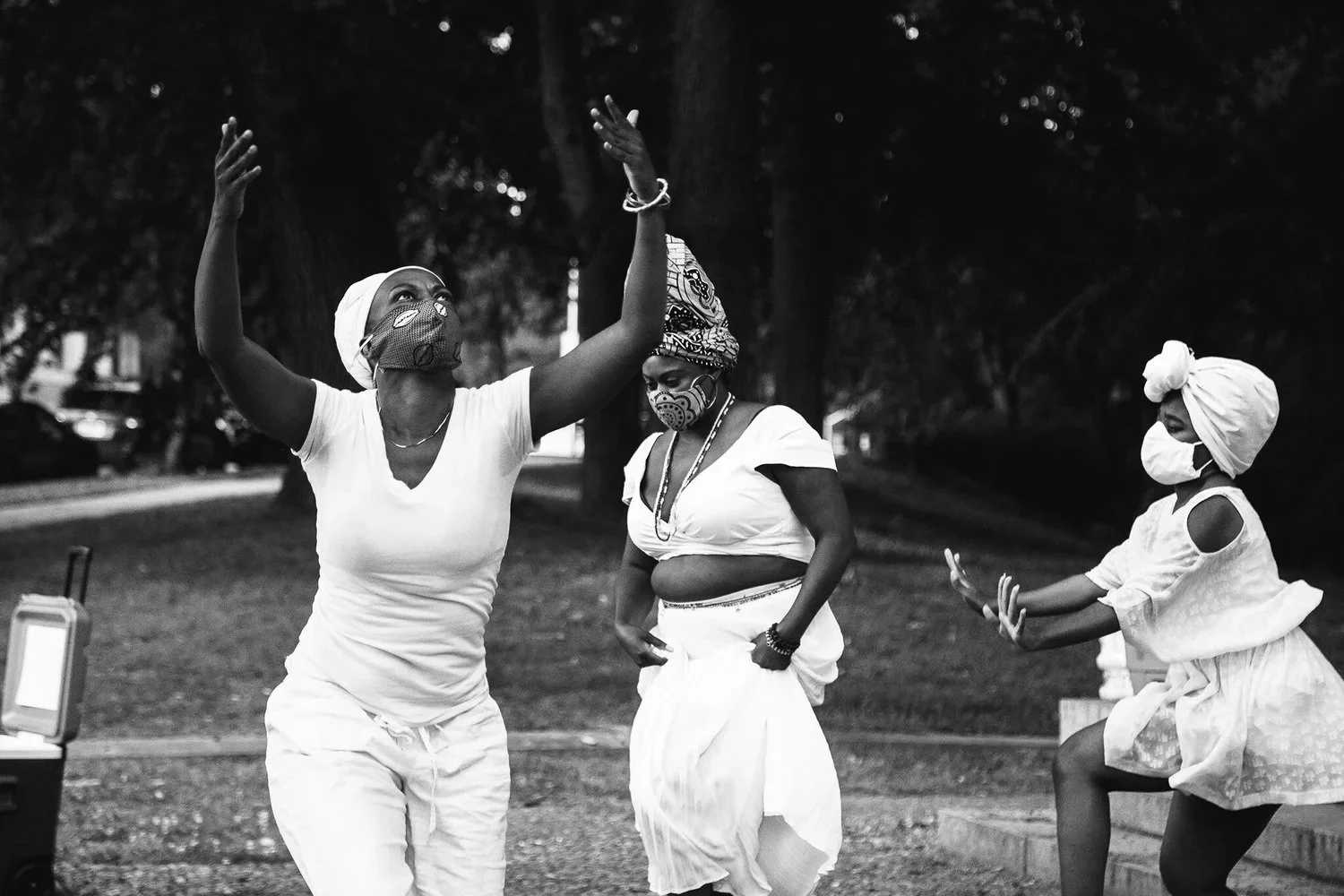 Three women dancing outdoors, wearing headwraps and face masks, with trees and parked cars in the background.