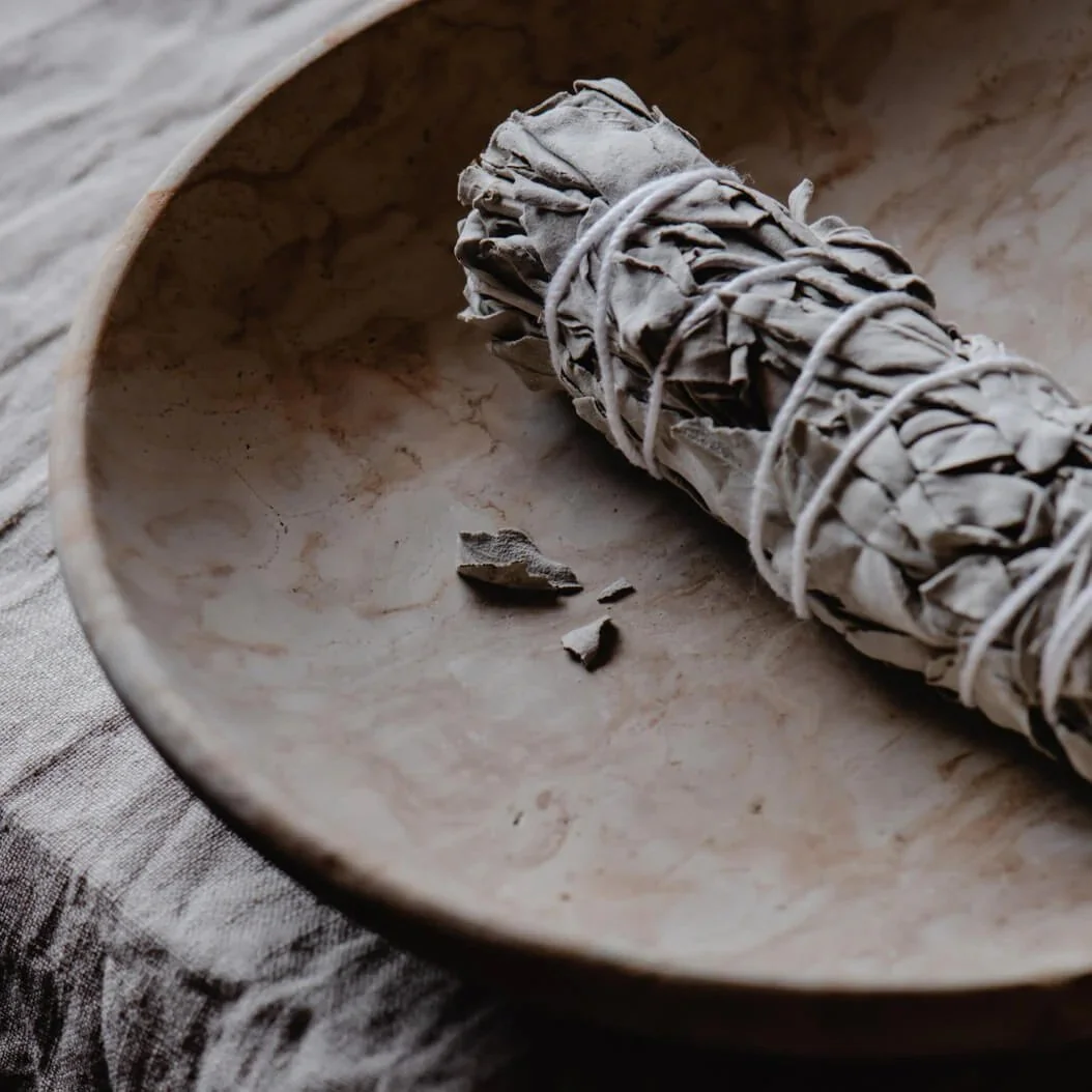 A bundle of sage, tied with string, resting inside a marble bowl with some sage leaves near the bundle.