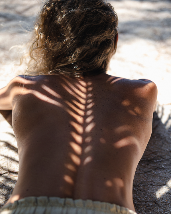 Back of a woman lying on sand with sunlight creating a shadow pattern resembling a leaf or feather on her back.