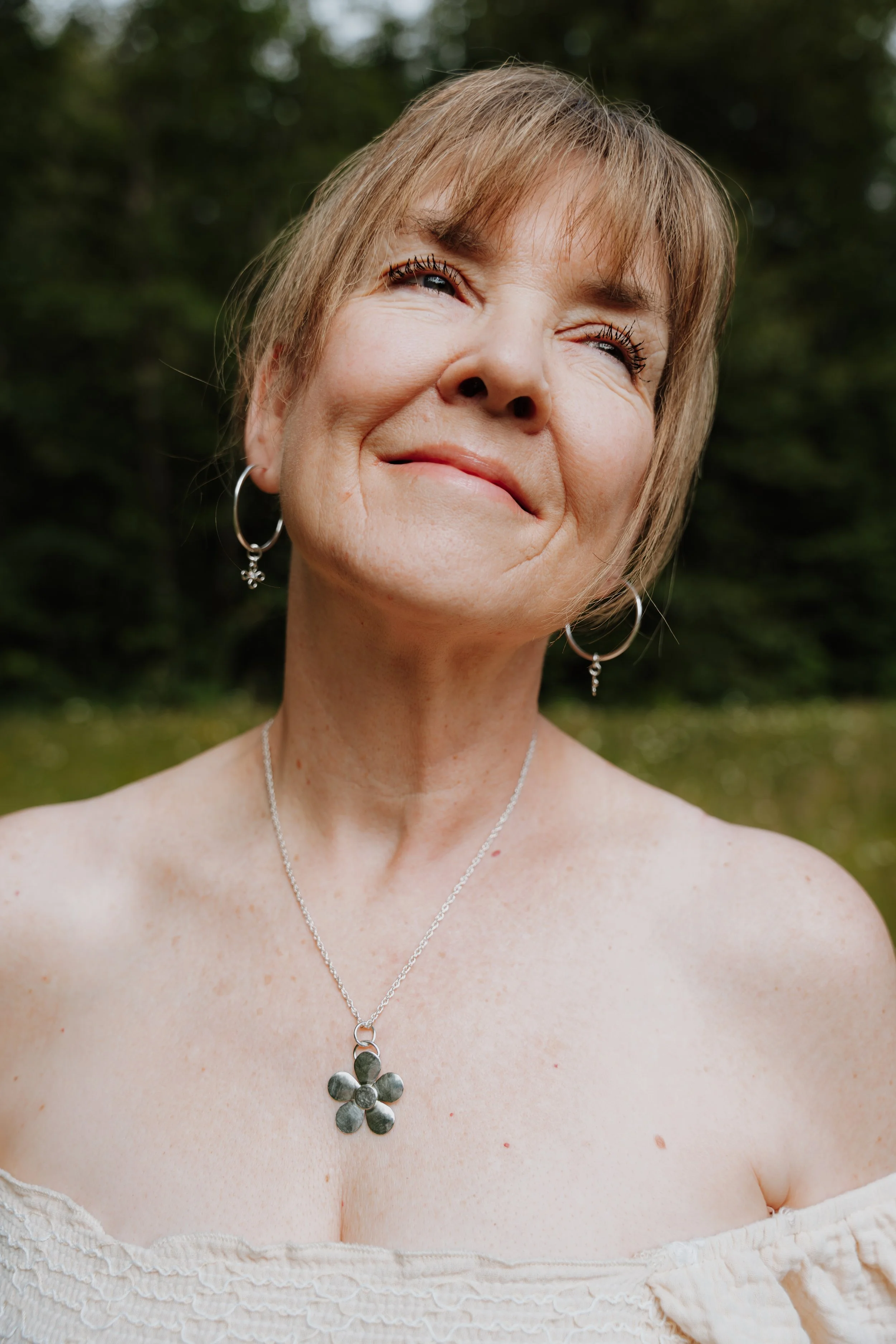 Close-up of a smiling woman with short blond hair, wearing earrings and a necklace, outdoors with a blurred green background.