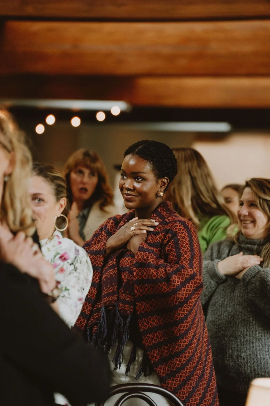 A woman with short curly black hair wearing a red and black patterned shawl hears the speaker with her hands placed over her chest in a group of women at an indoor event.
