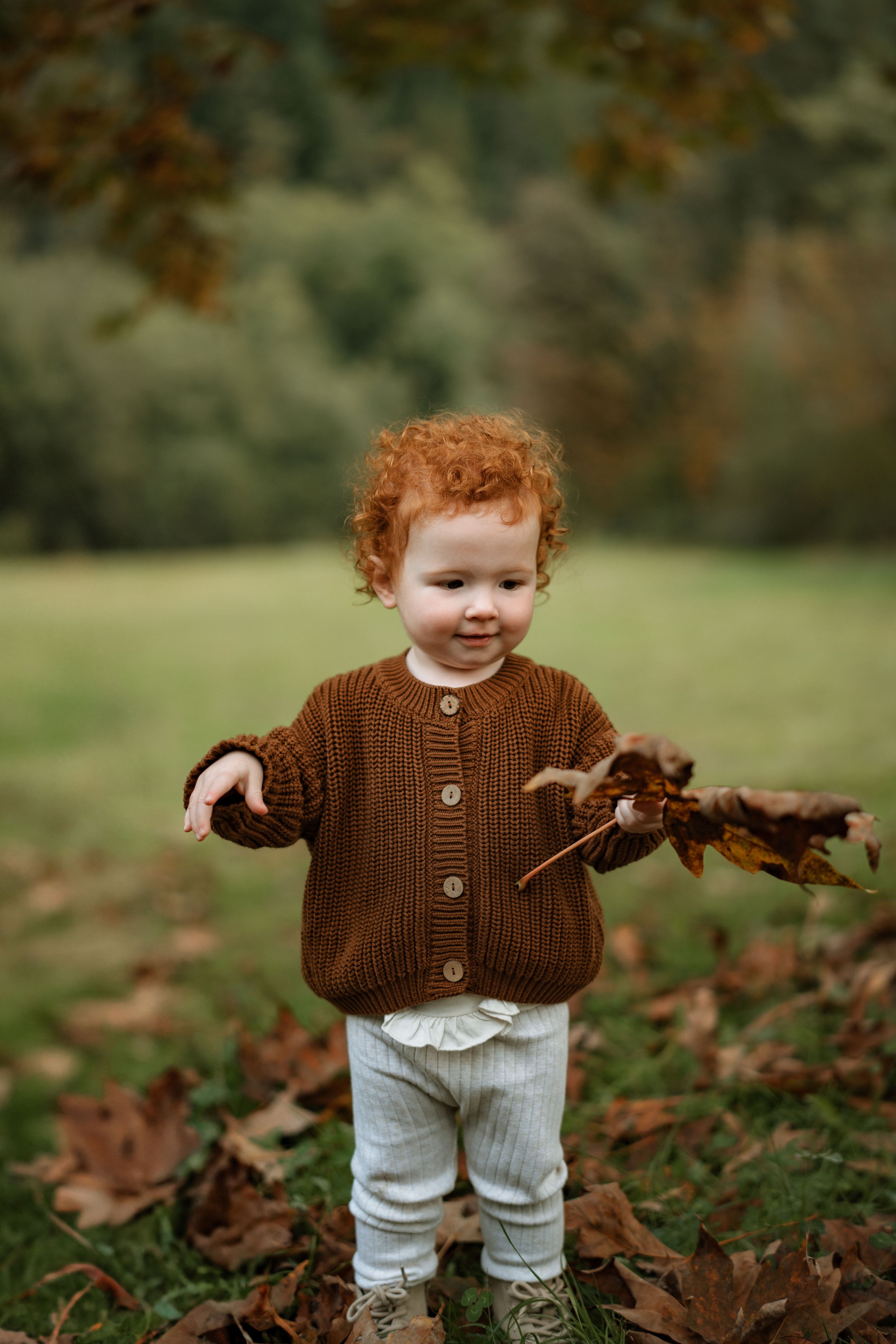 A young child with red curly hair wearing a brown cardigan and beige pants, playing with fallen leaves outdoors in a park or forest during autumn.