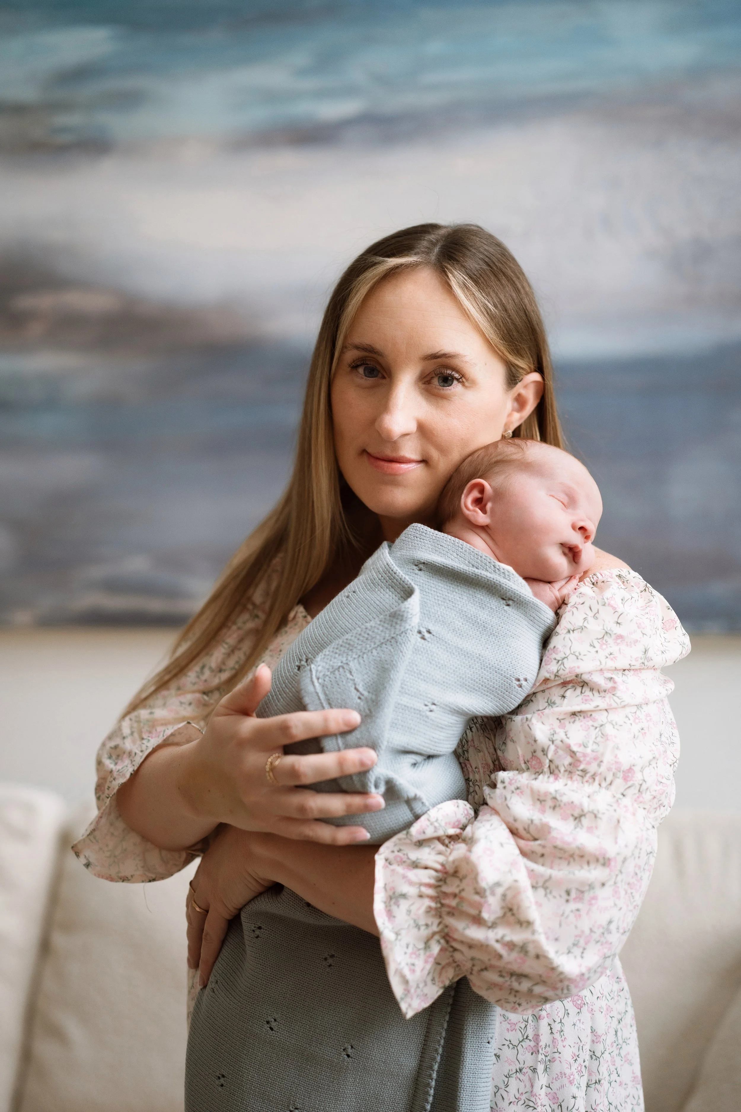 A woman with long blonde hair holding a sleeping baby in her arms. The background features a painting of a cloudy sky over the ocean.