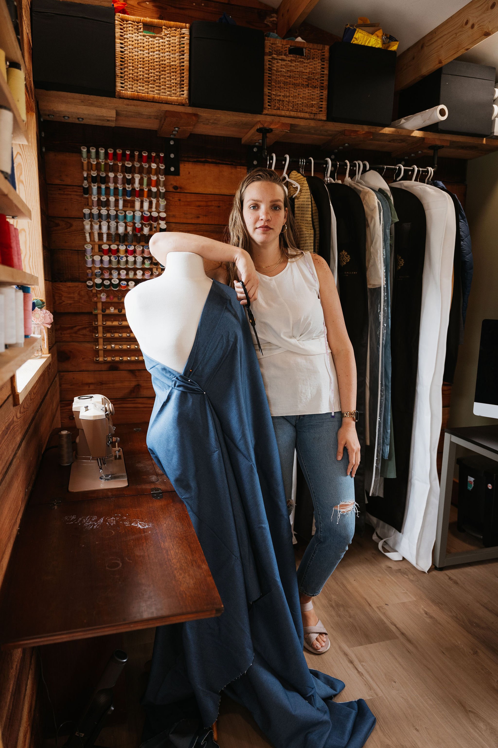 A woman standing in a room surrounded by clothing and sewing supplies. She has long hair, wearing a white sleeveless top and ripped jeans, leaning on a mannequin with a dress. Behind her are garment bags and a wall with a spice rack and storage boxes